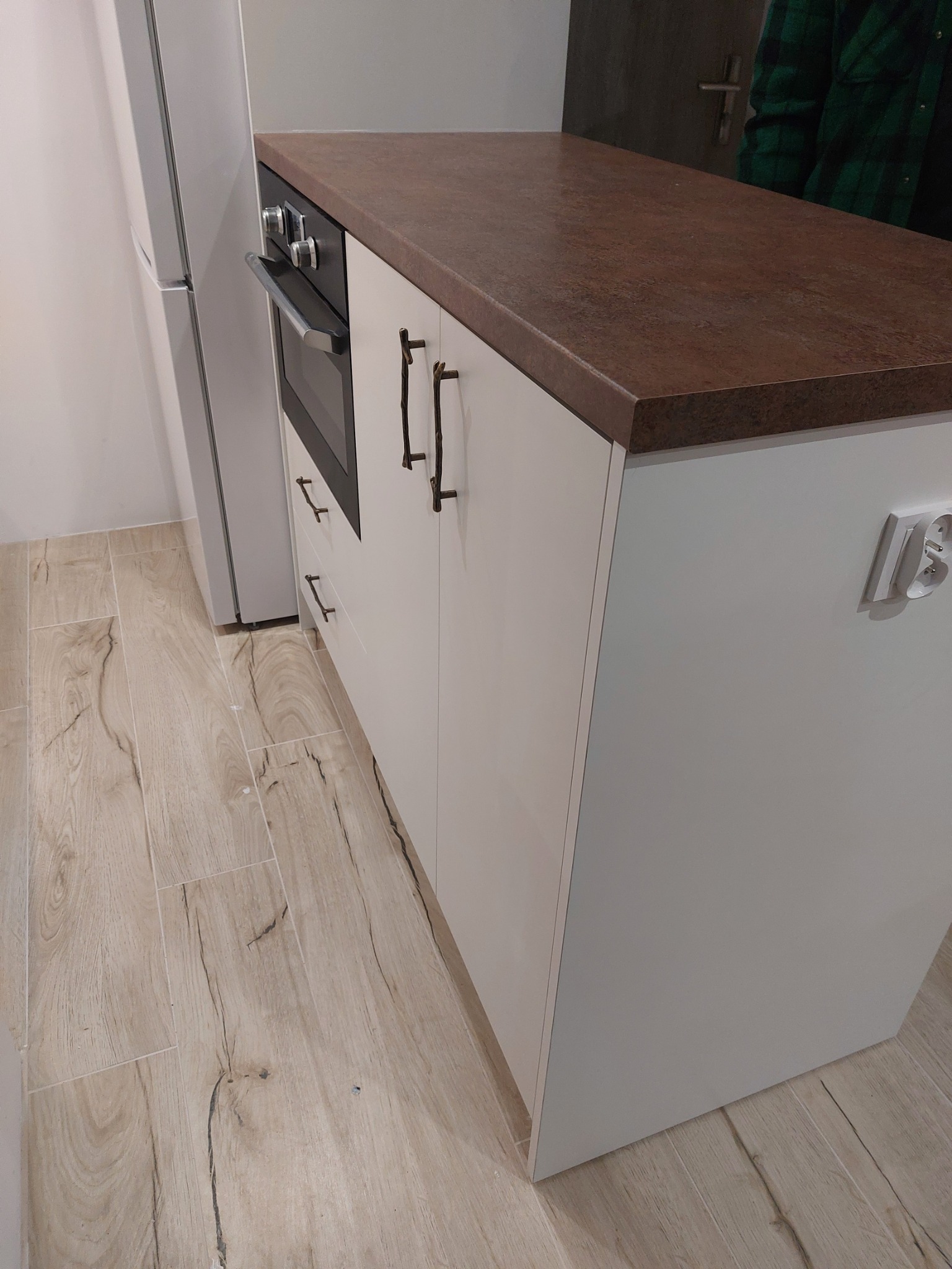 Fragment kuchni z białymi szafkami, brązowym blatem i piekarnikiem, widoczne drewnopodobne płytki podłogowe i gniazdko elektryczne na ścianie.