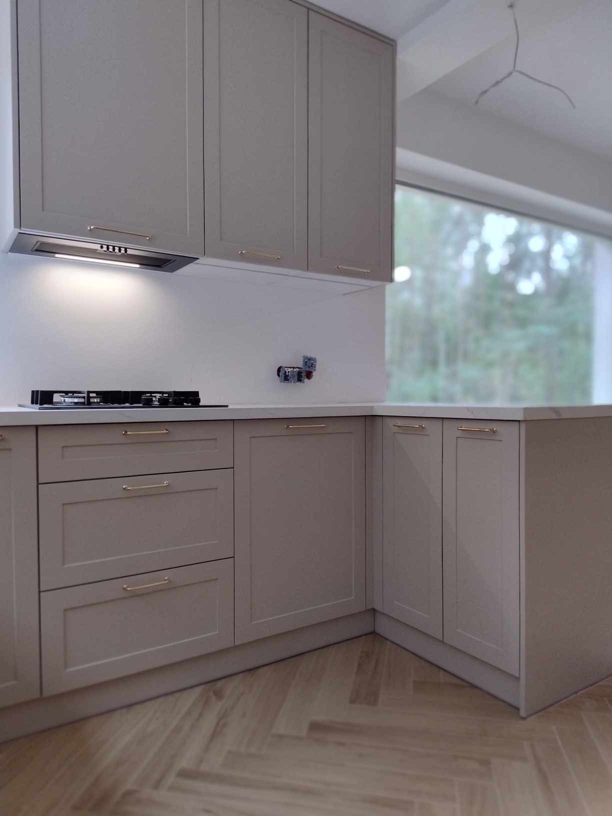 Jasnoszara zabudowa kuchenna z frezowanymi frontami i złotymi uchwytami, widoczna płyta gazowa i okap, parkiet w jodełkę, Warszawa.