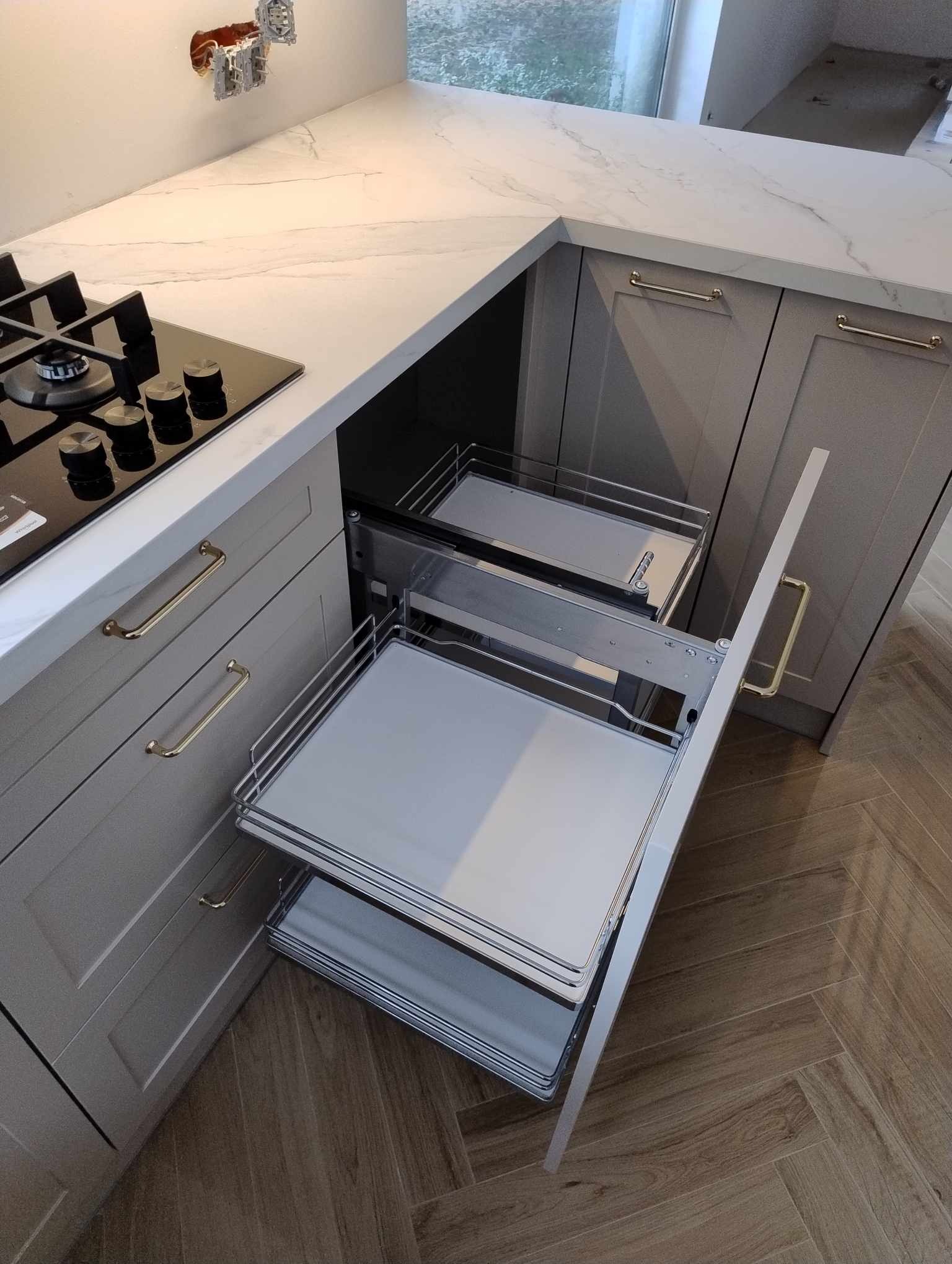 Szare szafki kuchenne z wysuwanymi koszami cargo, złote uchwyty, blat imitujący marmur, płyta indukcyjna i parkiet w jodełkę.