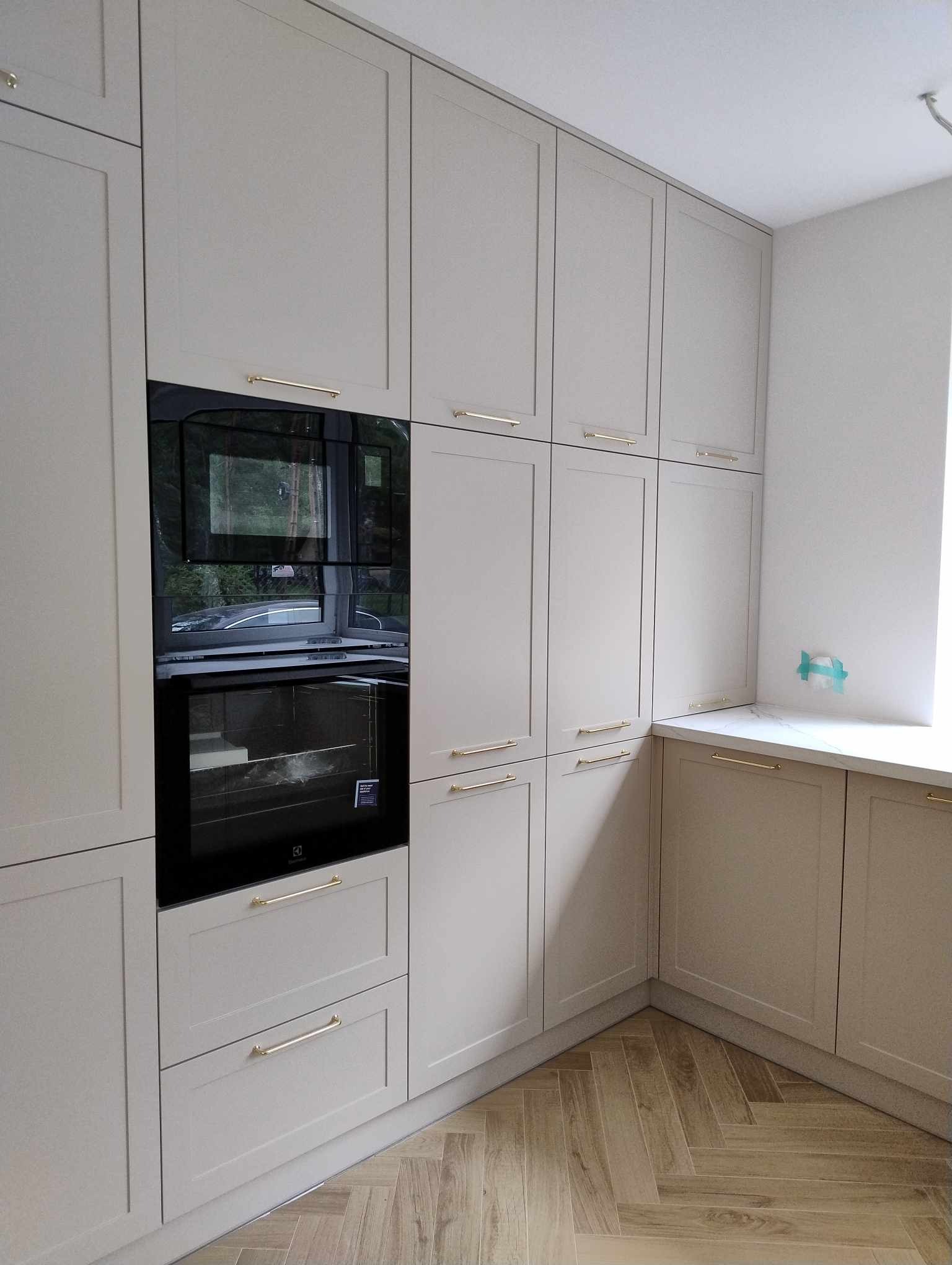 Jasnoszara zabudowa kuchenna z piekarnikiem w zabudowie i złotymi uchwytami, parkiet ułożony w jodełkę.