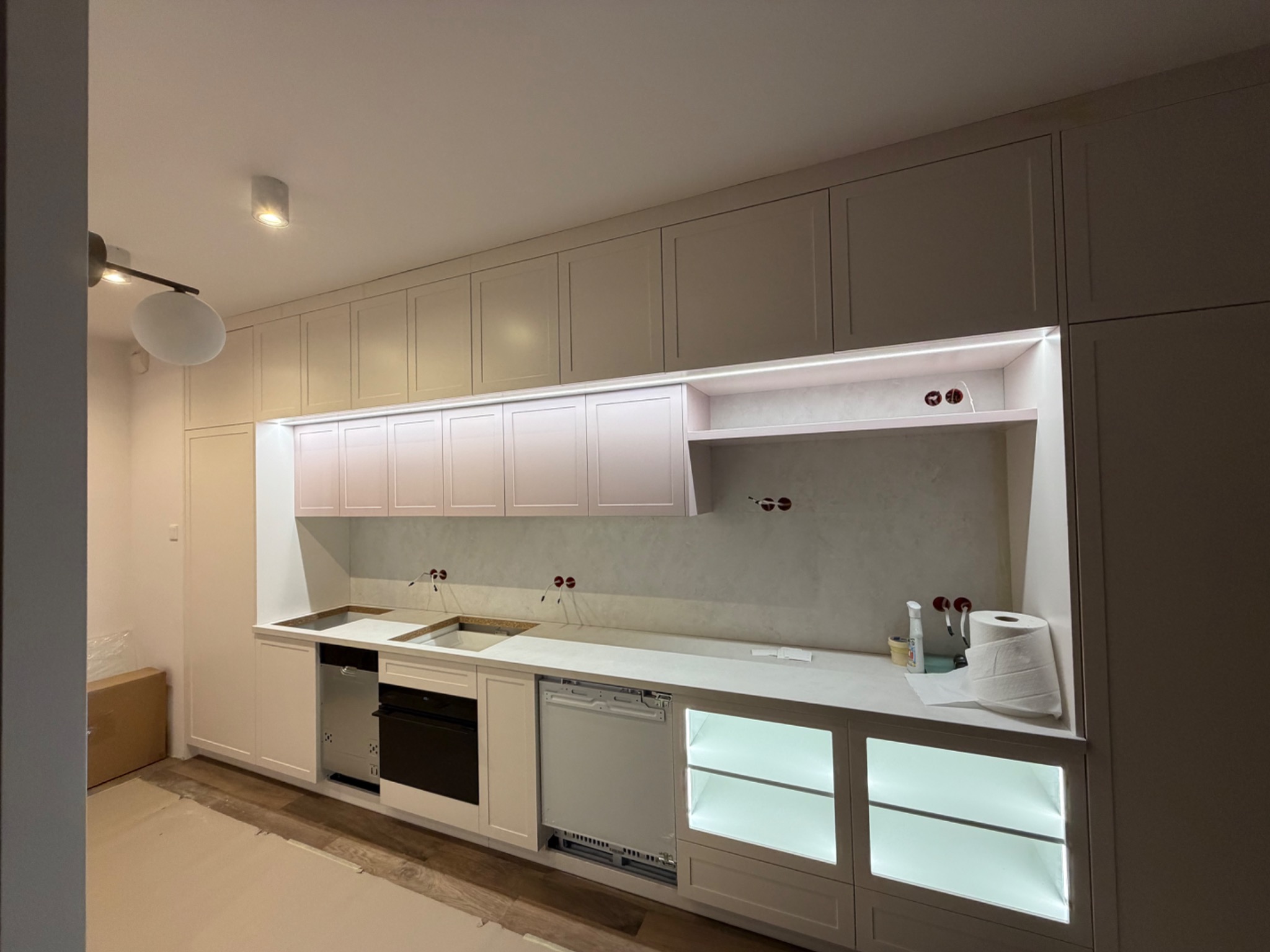 Jasna kuchnia z oświetleniem LED pod szafkami, widoczne przygotowane instalacje wodne i elektryczne. Częściowo widoczne kartony na podłodze.