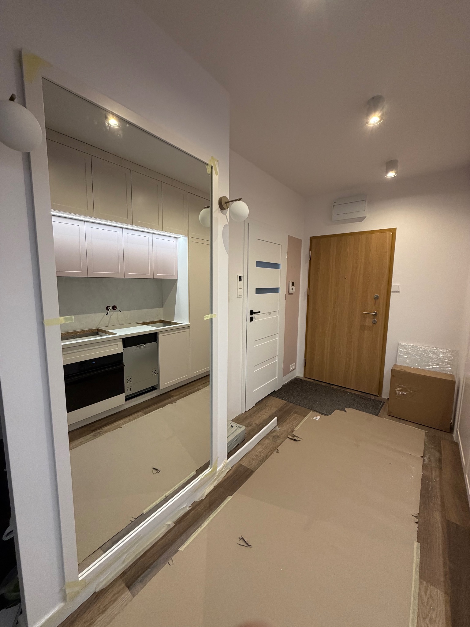 Nowoczesna kuchnia z jasnymi szafkami, widziana w lustrzanym odbiciu w przedpokoju. Widoczne zabezpieczenia podłogi kartonem. Minimalistyczny design.
