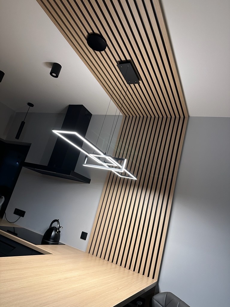 Nowoczesna kuchnia z drewnianymi lamelami na suficie i ścianie, oświetleniem LED i czarnym okapem. Jasny blat roboczy i minimalistyczny design.
