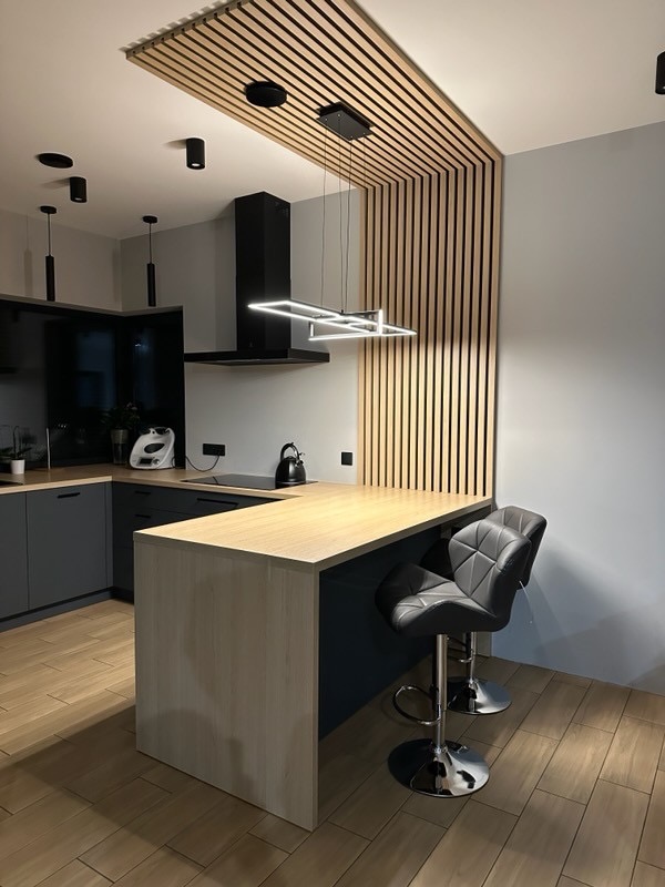 Nowoczesna kuchnia z drewnianą zabudową sufitową i wyspą z blatem w kolorze jasnego drewna. Czarne szafki i oświetlenie punktowe. Dwa hokery przy wyspie.