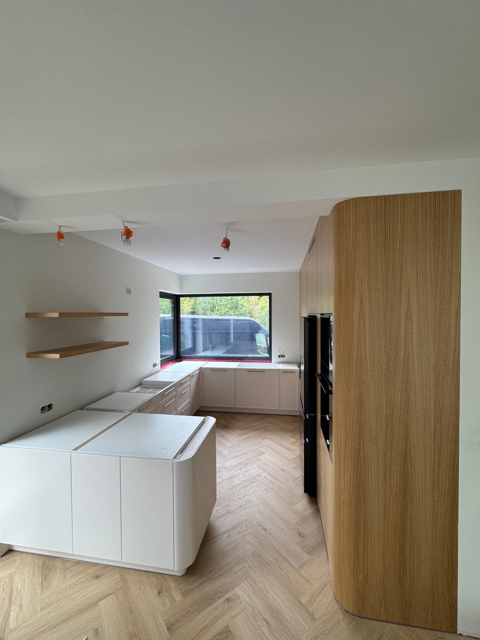 Jasna kuchnia w trakcie montażu: białe szafki z zaokrąglonymi frontami, drewniane półki i parkiet w jodełkę. Widoczne niedokończone elementy i oświetlenie budowlane.