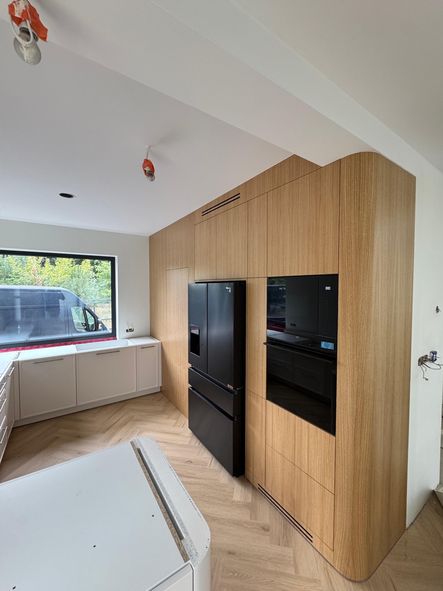 Nowoczesna kuchnia w trakcie montażu: drewniane fronty, czarna lodówka i piekarnik w zabudowie. Widoczny fragment blatu i okno z widokiem na zieleń.