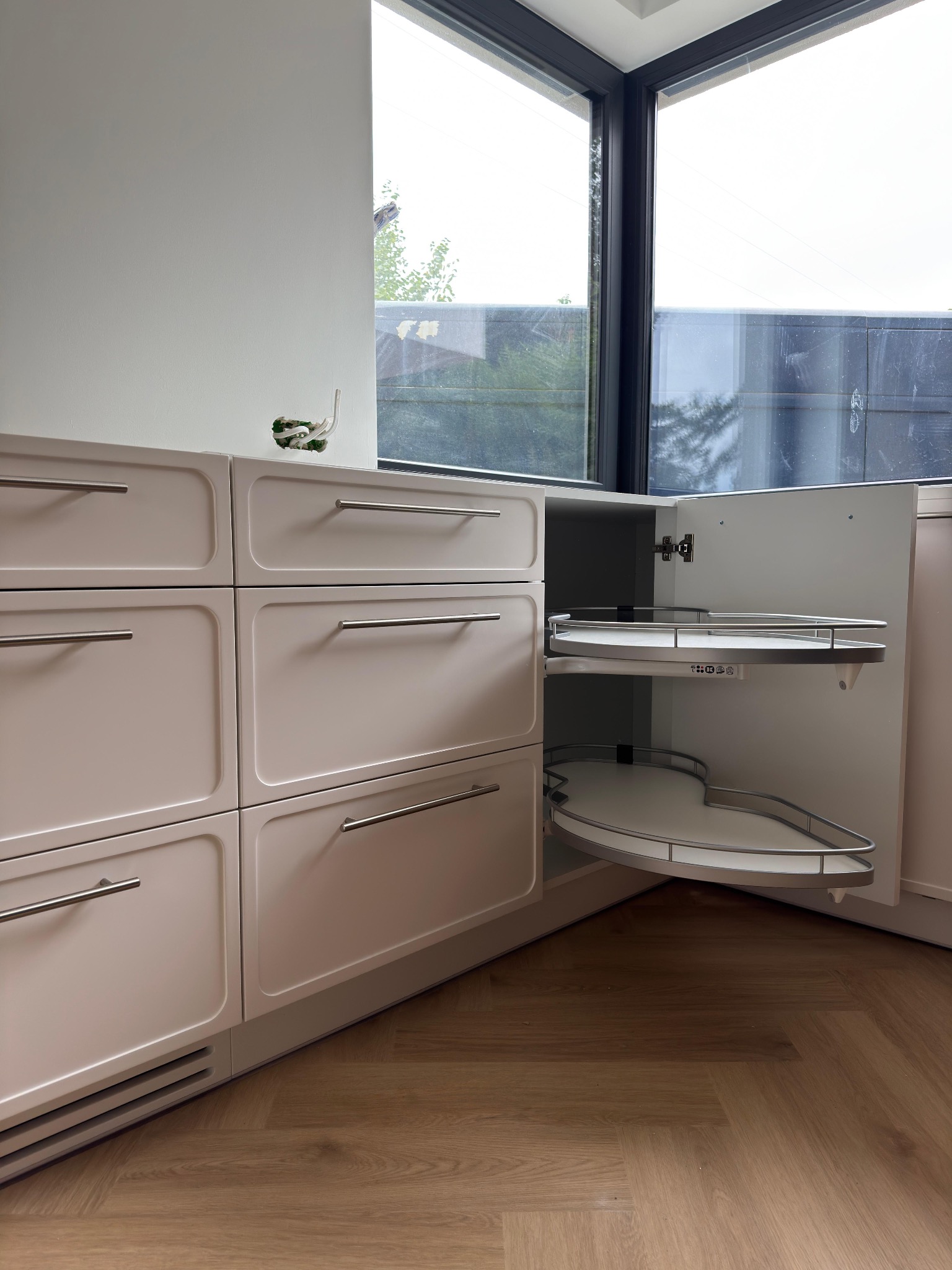 Jasna zabudowa kuchenna z otwartą szafką narożną z półkami obrotowymi, widoczna instalacja elektryczna nad blatem. Nowoczesny design, drewniana podłoga.