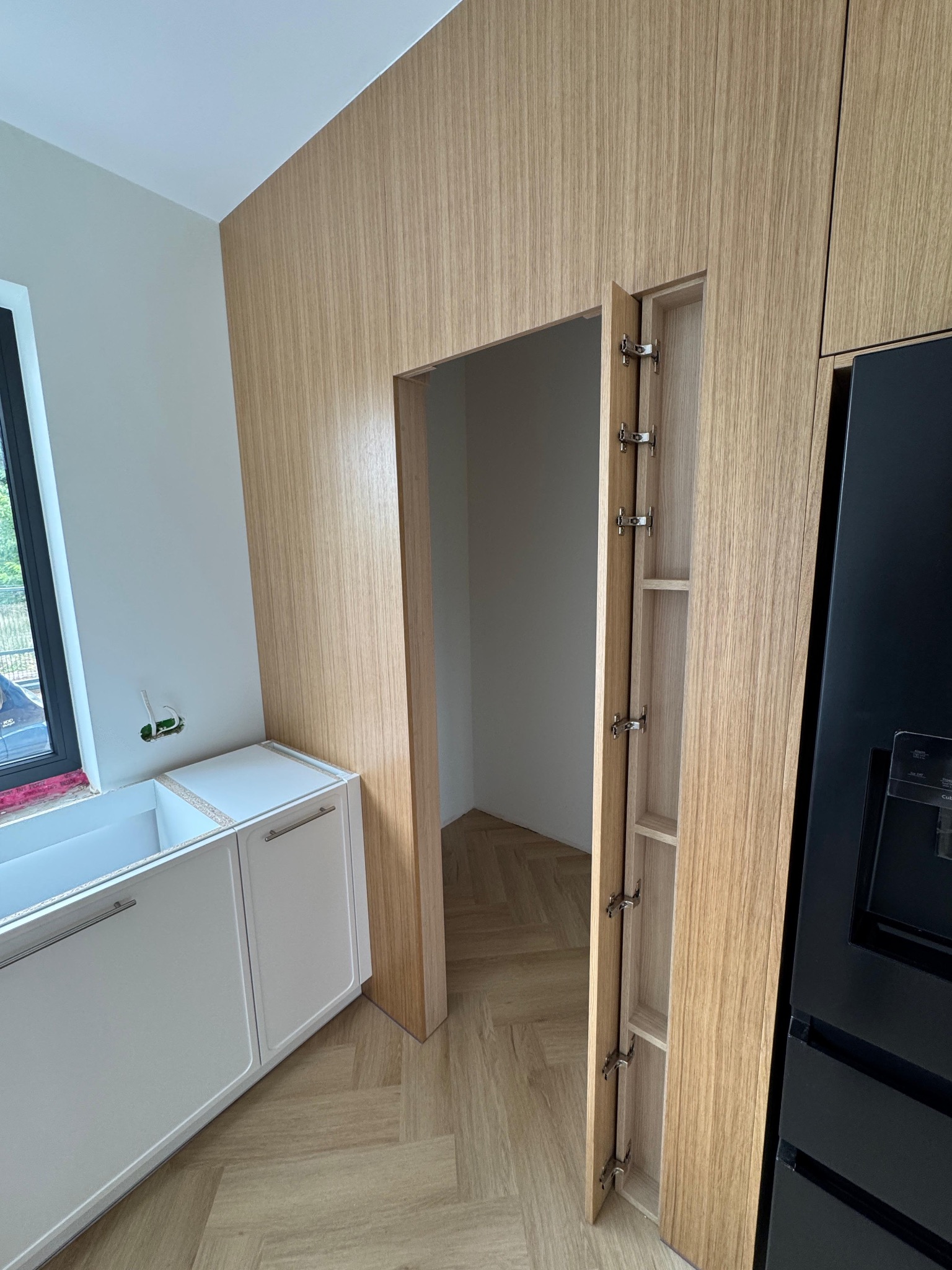 Nowoczesna kuchnia z drewnianą zabudową, otwartą szafką typu cargo i białymi szafkami dolnymi. Widoczny fragment czarnej lodówki i parkiet w jodełkę.