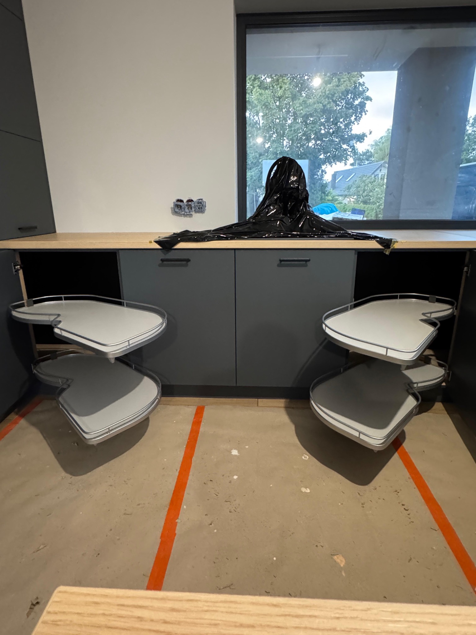 Szare szafki kuchenne z wysuwanymi półkami narożnymi, zabezpieczone folią. Widoczne gniazdka elektryczne i okno z widokiem na zieleń. Podłoga z pomarańczowymi liniami.