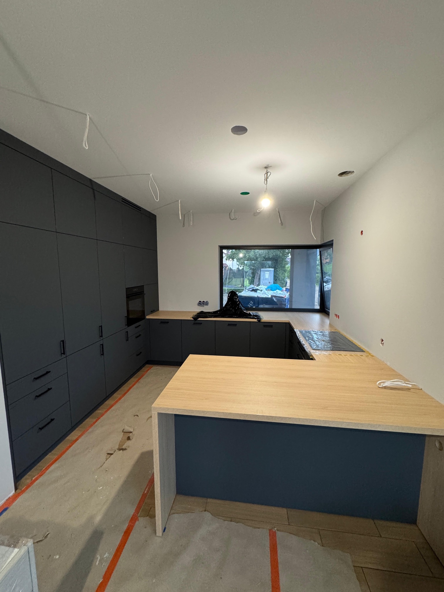 Ciemnoszara zabudowa kuchenna z drewnianym blatem w surowym wnętrzu. Widoczne niedokończone wykończenia podłogi i wiszące przewody elektryczne.