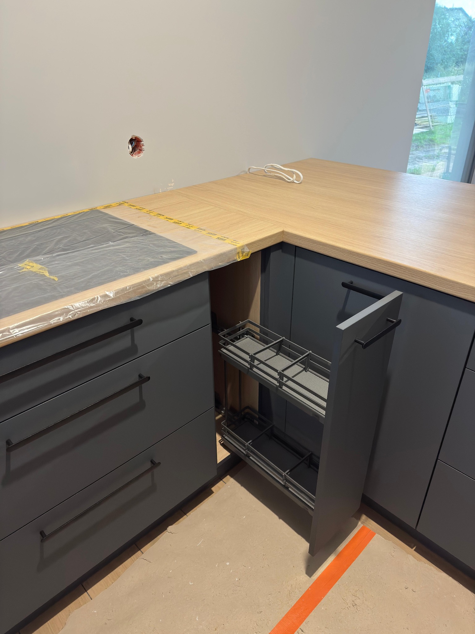 Szara zabudowa kuchenna z drewnianym blatem i wysuwanym koszem cargo. Elementy zabezpieczone folią. Widoczny fragment okna i niedokończone wykończenie podłogi.