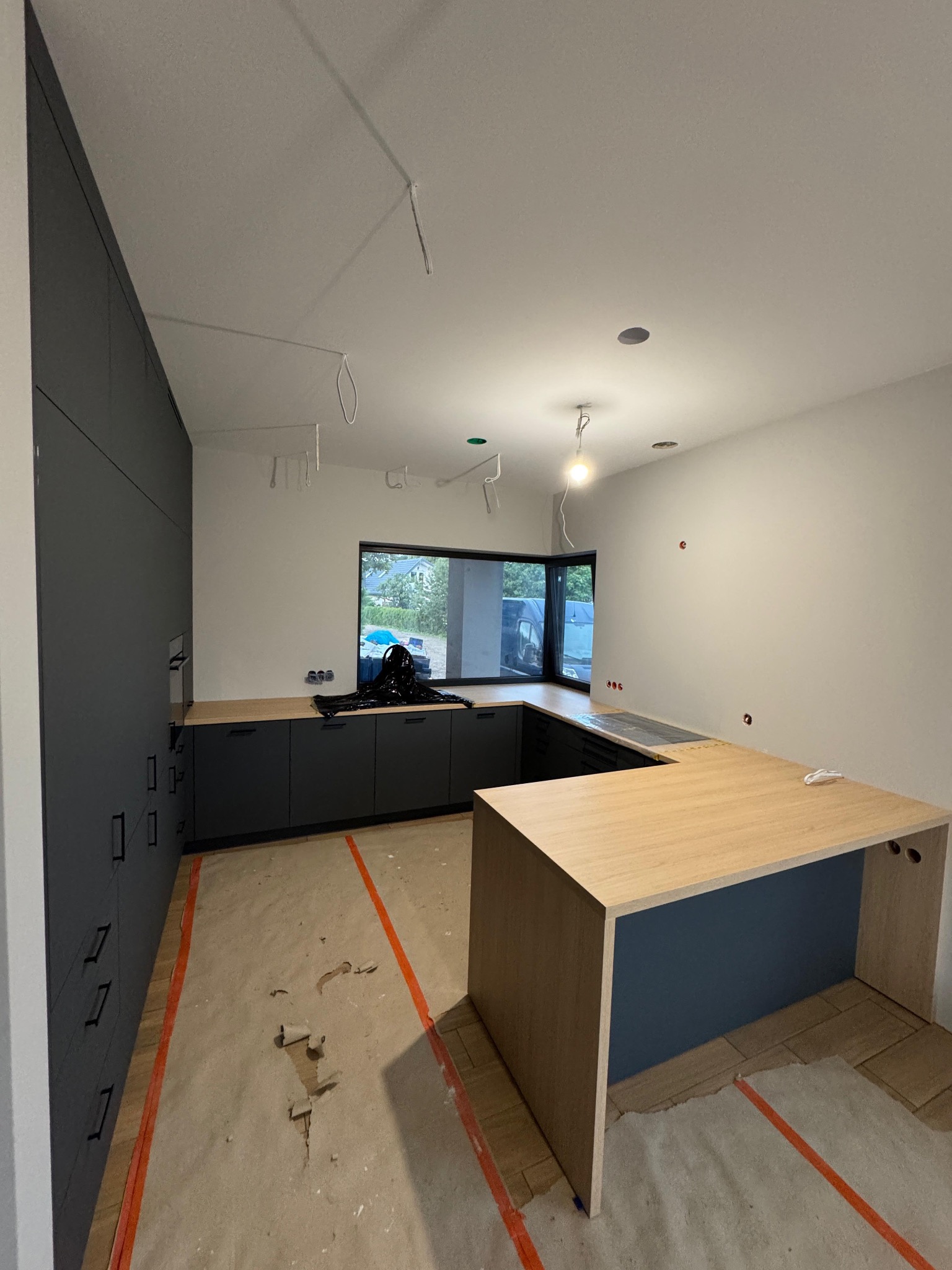 Nowoczesna kuchnia w trakcie montażu: grafitowe szafki, jasny blat, widoczne zabezpieczenia podłogi. Minimalistyczny design, surowe wykończenie, okno z widokiem na zewnątrz.