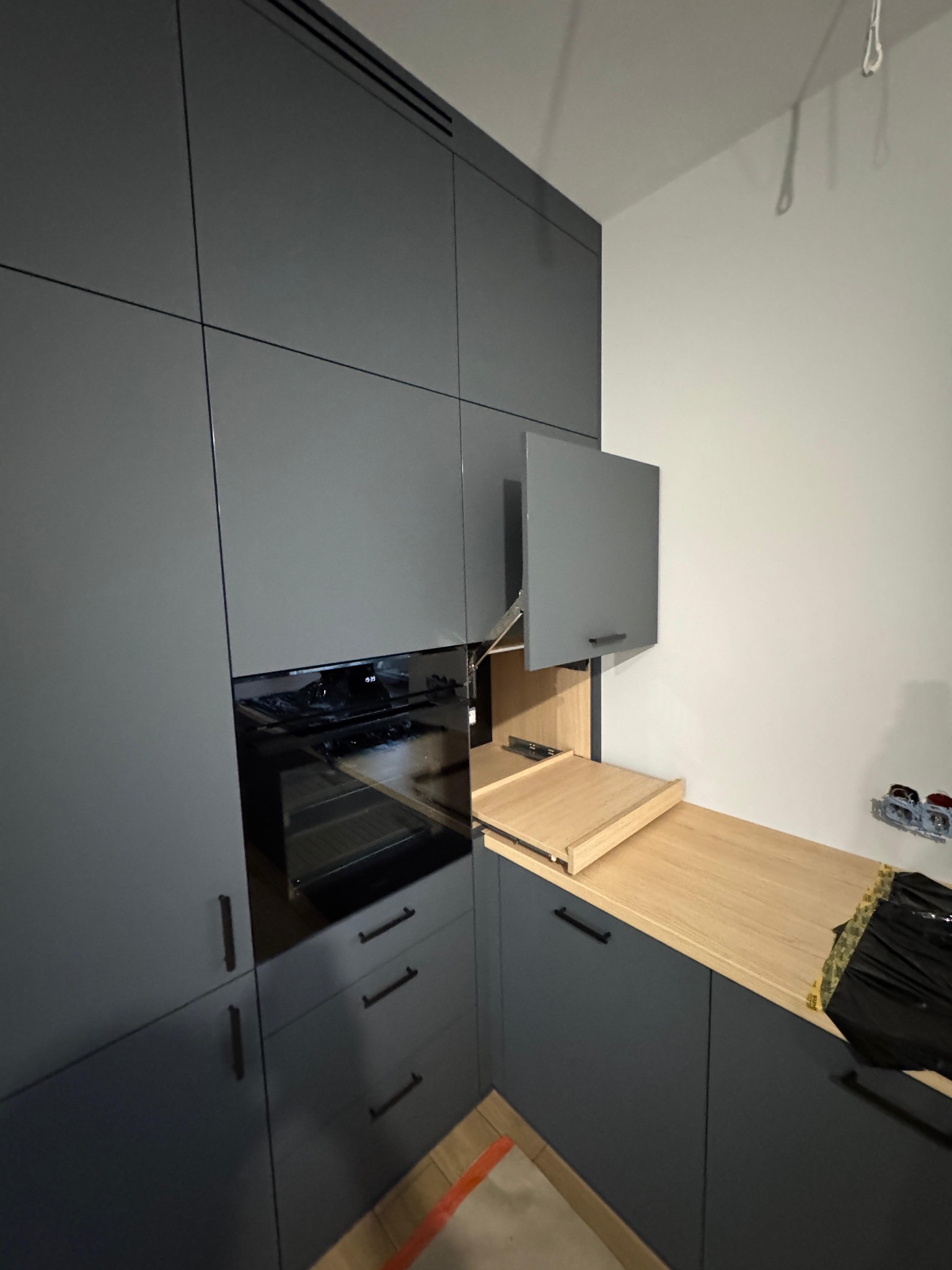 Szara zabudowa kuchenna z piekarnikiem i drewnianym blatem, szafka uchylna otwarta. Minimalistyczny design, czarne uchwyty, widoczne gniazdka elektryczne w tle.