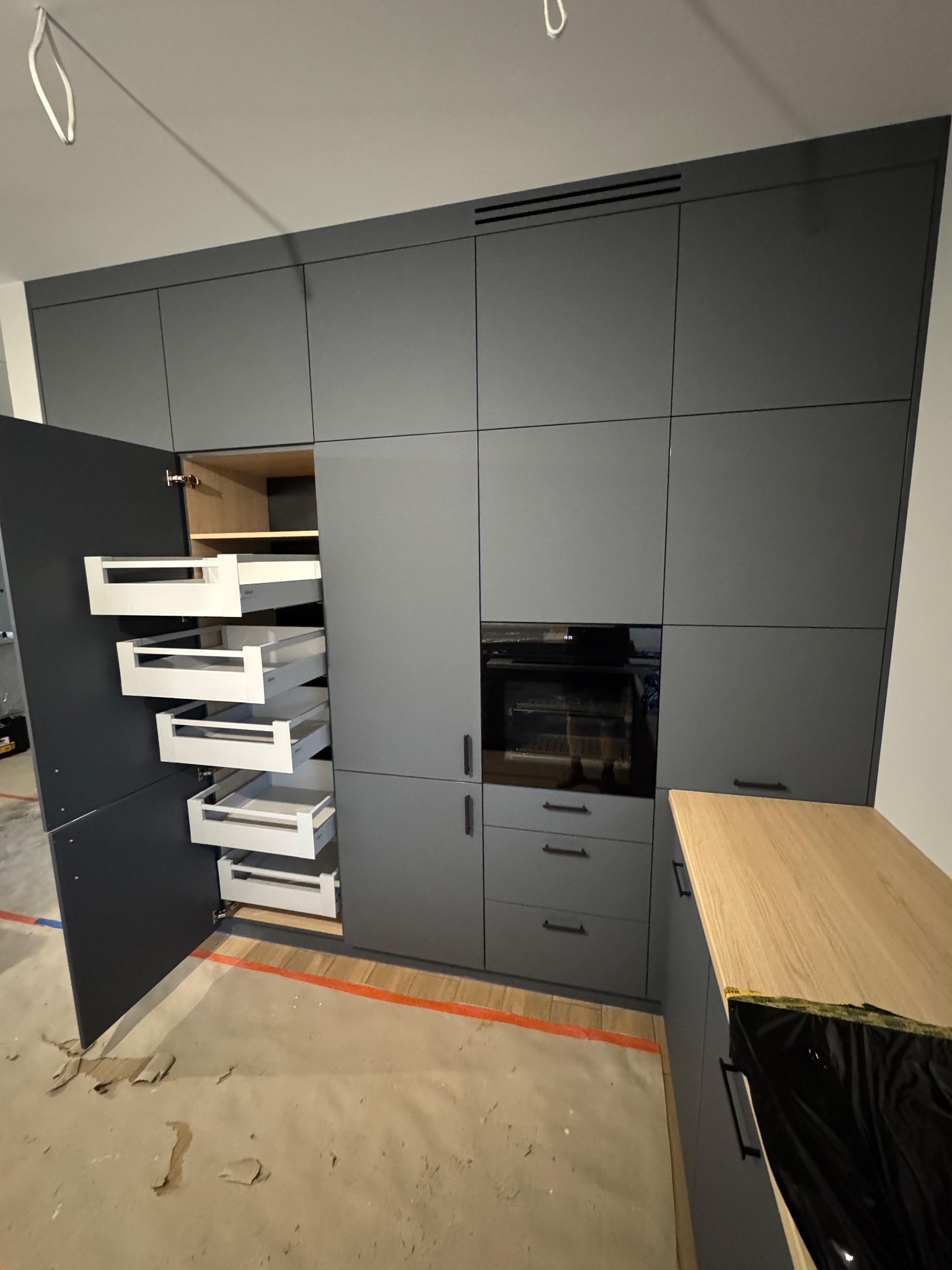 Szara zabudowa kuchenna z otwartą szafką cargo, ukazującą wysuwane, białe półki. Widoczny fragment blatu drewnianego i sprzęt AGD. Podłoga zabezpieczona folią malarską.