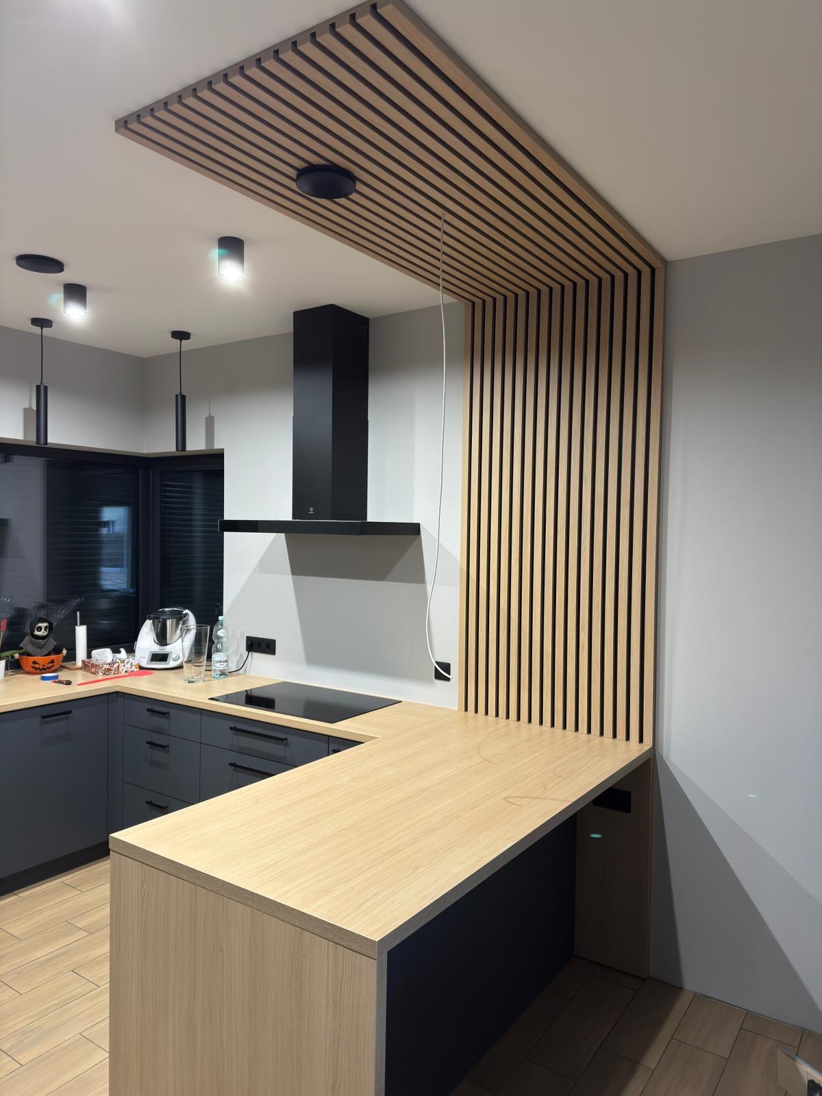 Nowoczesna kuchnia z drewnianymi lamelami na suficie i ścianie, szare szafki, jasny blat i czarny okap. Minimalistyczny design z akcentem naturalnego drewna.