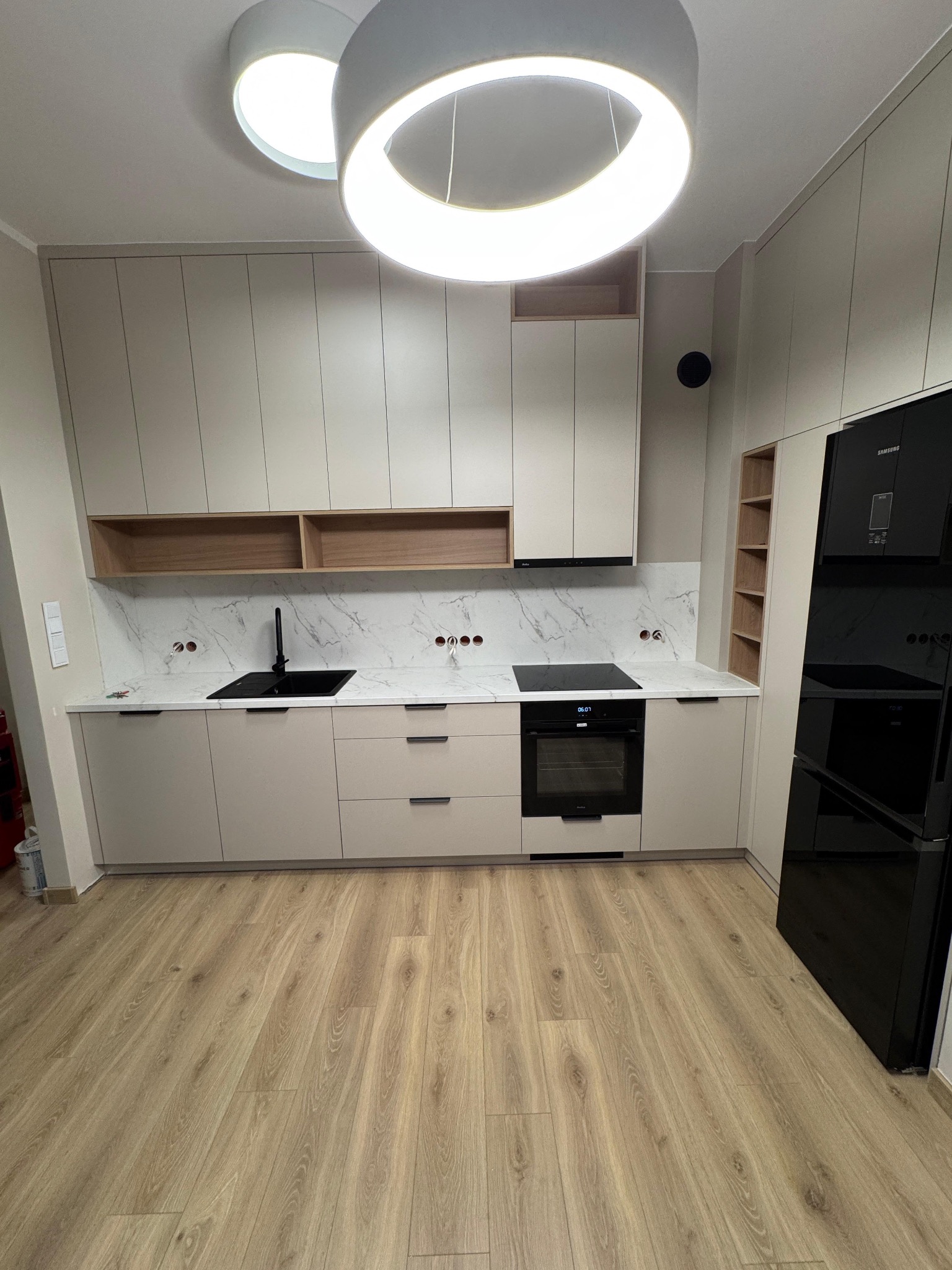 Jasna kuchnia z beżowymi szafkami, czarnym zlewem i sprzętem AGD. Blat imitujący marmur, drewniane półki i podłoga. Okrągła lampa sufitowa.