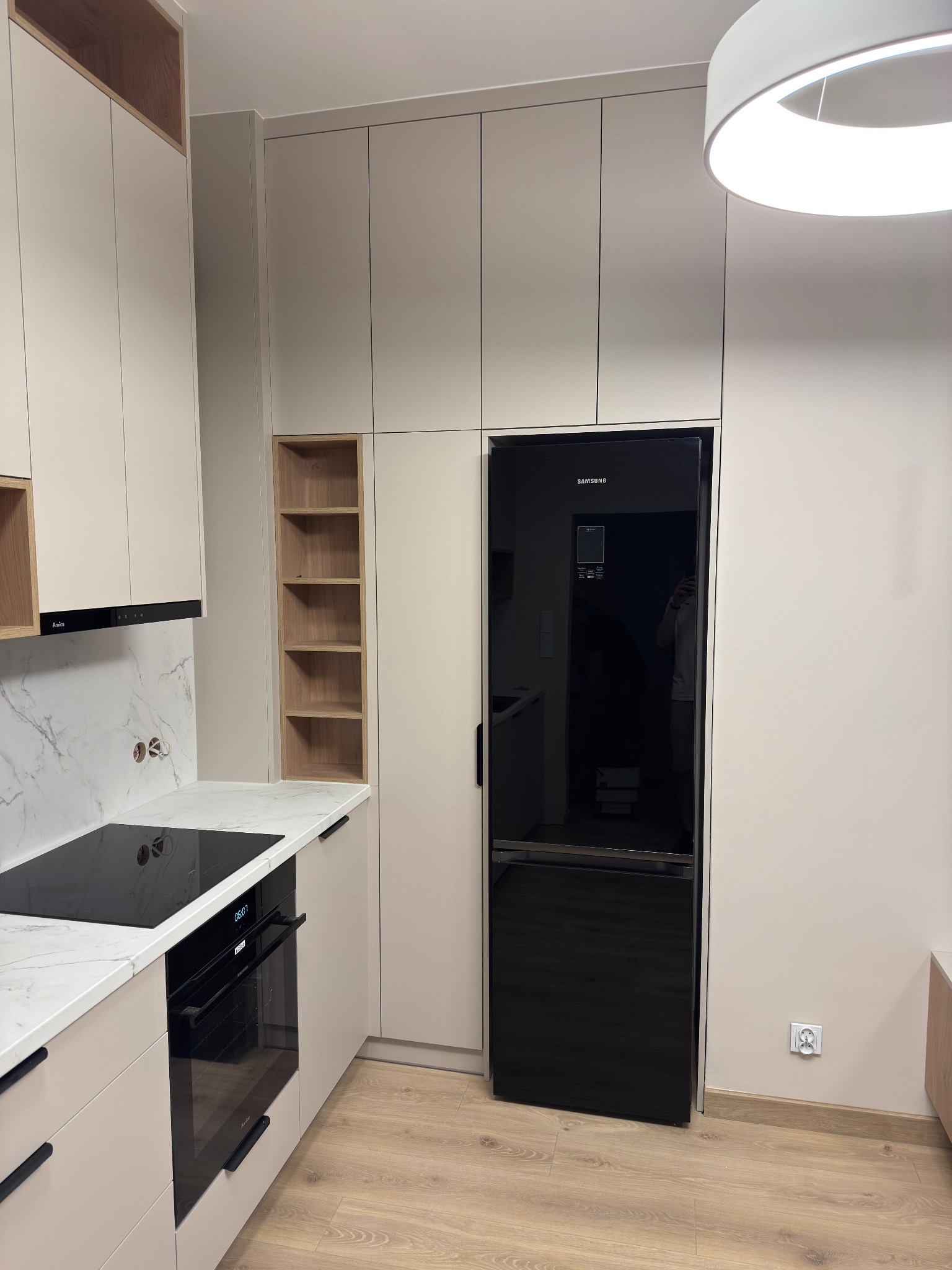 Beżowa zabudowa kuchenna z czarnym sprzętem AGD: piekarnik, płyta indukcyjna i lodówka Samsung. Marmurowy blat i drewniane półki w otwartej szafce.