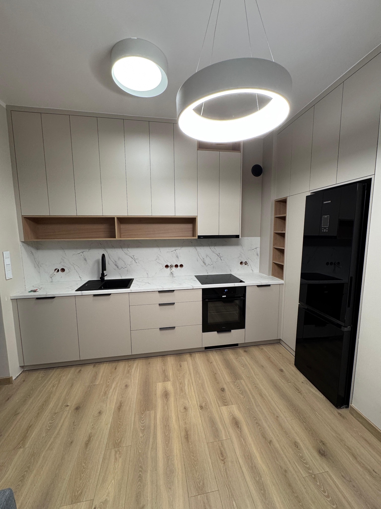 Jasna kuchnia z beżowymi szafkami, czarnym zlewem i sprzętem AGD. Marmurowy backsplash i drewniane półki dodają elegancji. Oświetlenie LED podkreśla nowoczesny design.