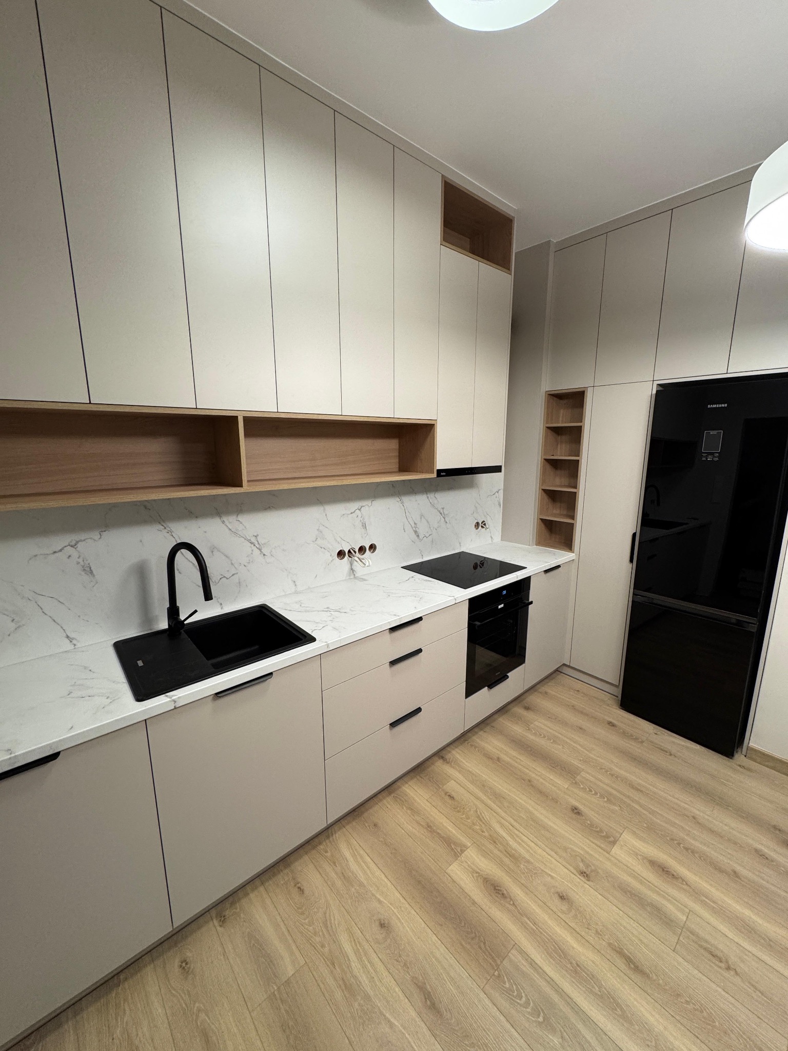 Jasna kuchnia z drewnianymi akcentami: szafki górne i dolne w odcieniach beżu, marmurowy blat, czarny zlew i sprzęt AGD. Otwarta półka z jasnego drewna.