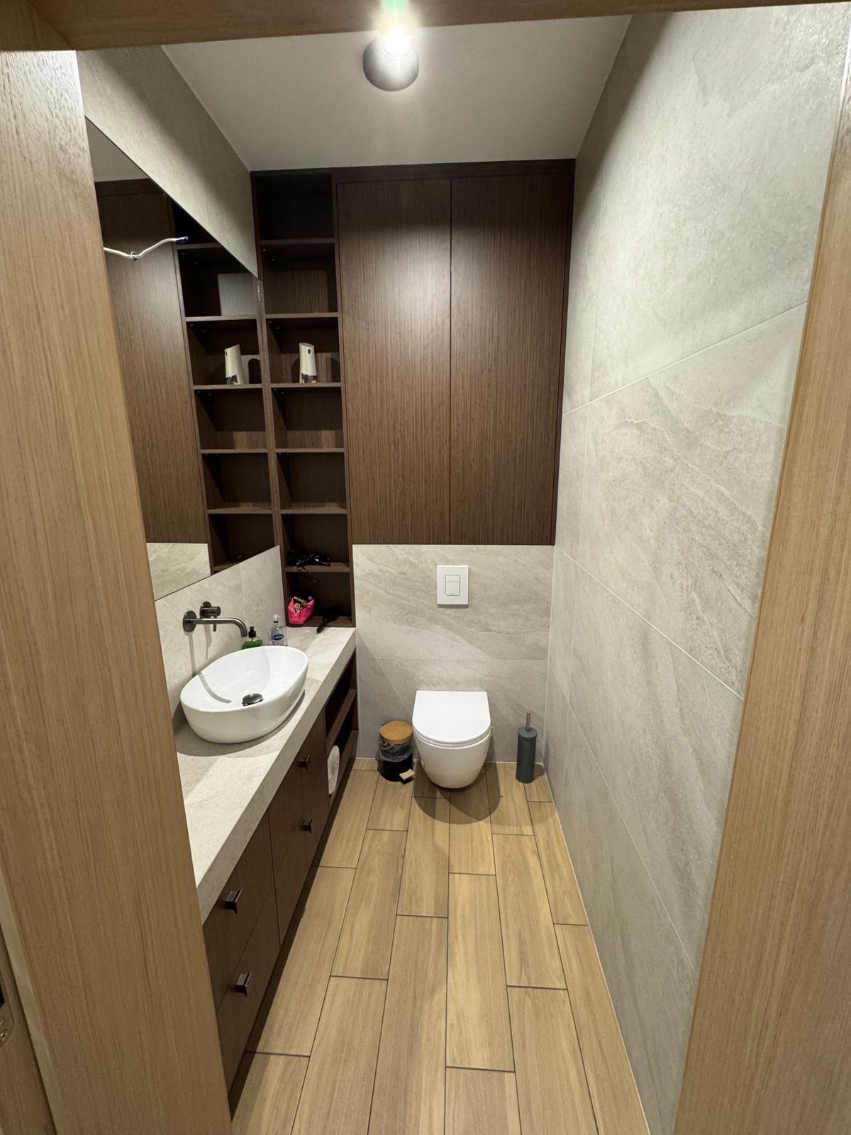 Nowoczesna łazienka w Warszawie: zabudowa z szafkami z ciemnego drewna nad umywalką z blatem z jasnego kamienia, toaleta pod ścianą z szarych płytek.