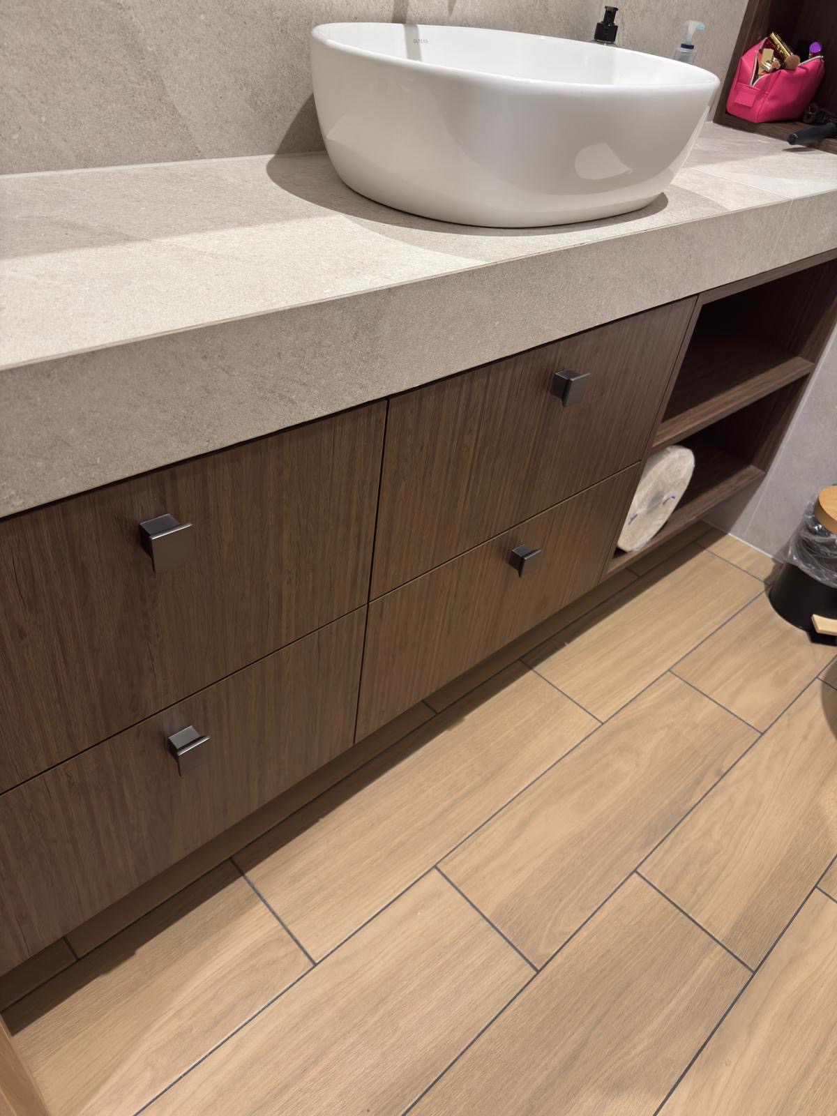 Nowoczesna łazienka z drewnianą szafką pod umywalką nablatową i blatem z szarego kamienia. Widoczne minimalistyczne uchwyty i drewniana podłoga.