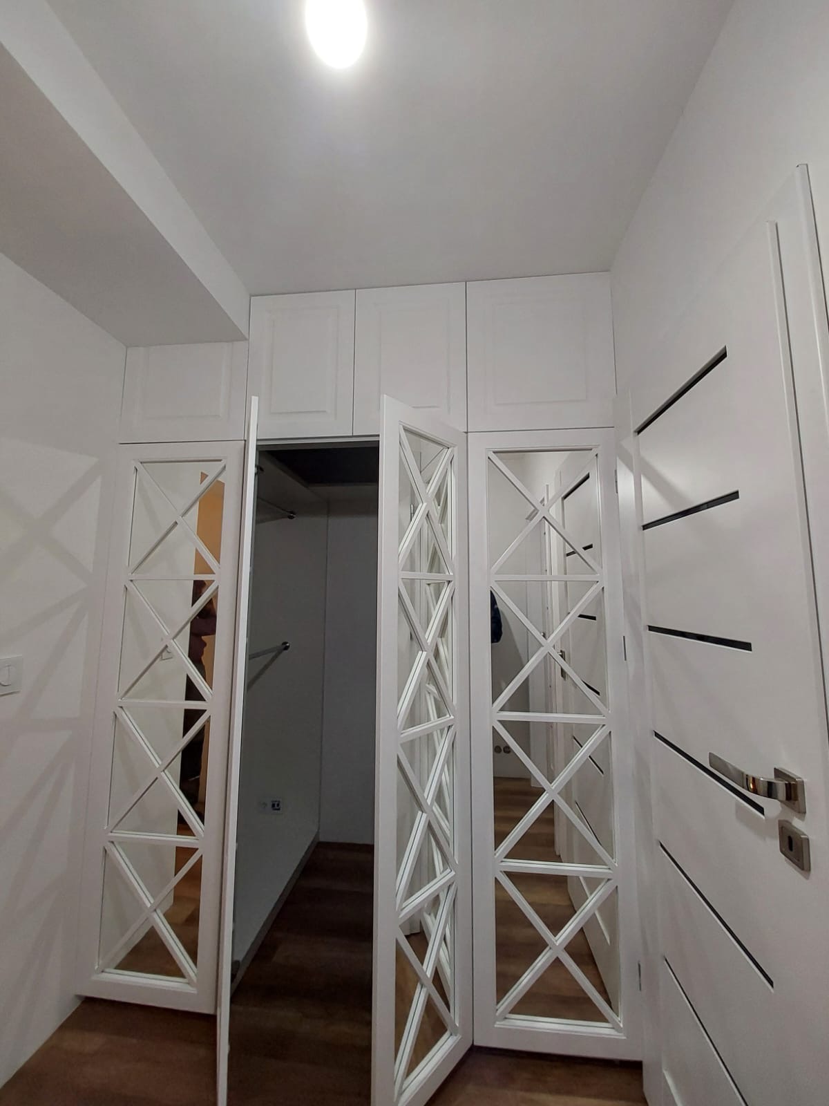 Biała szafa wnękowa z lustrzanymi drzwiami w geometryczne wzory, częściowo otwarta, ukazująca wnętrze garderoby. Widok z dołu, oświetlenie sufitowe.
