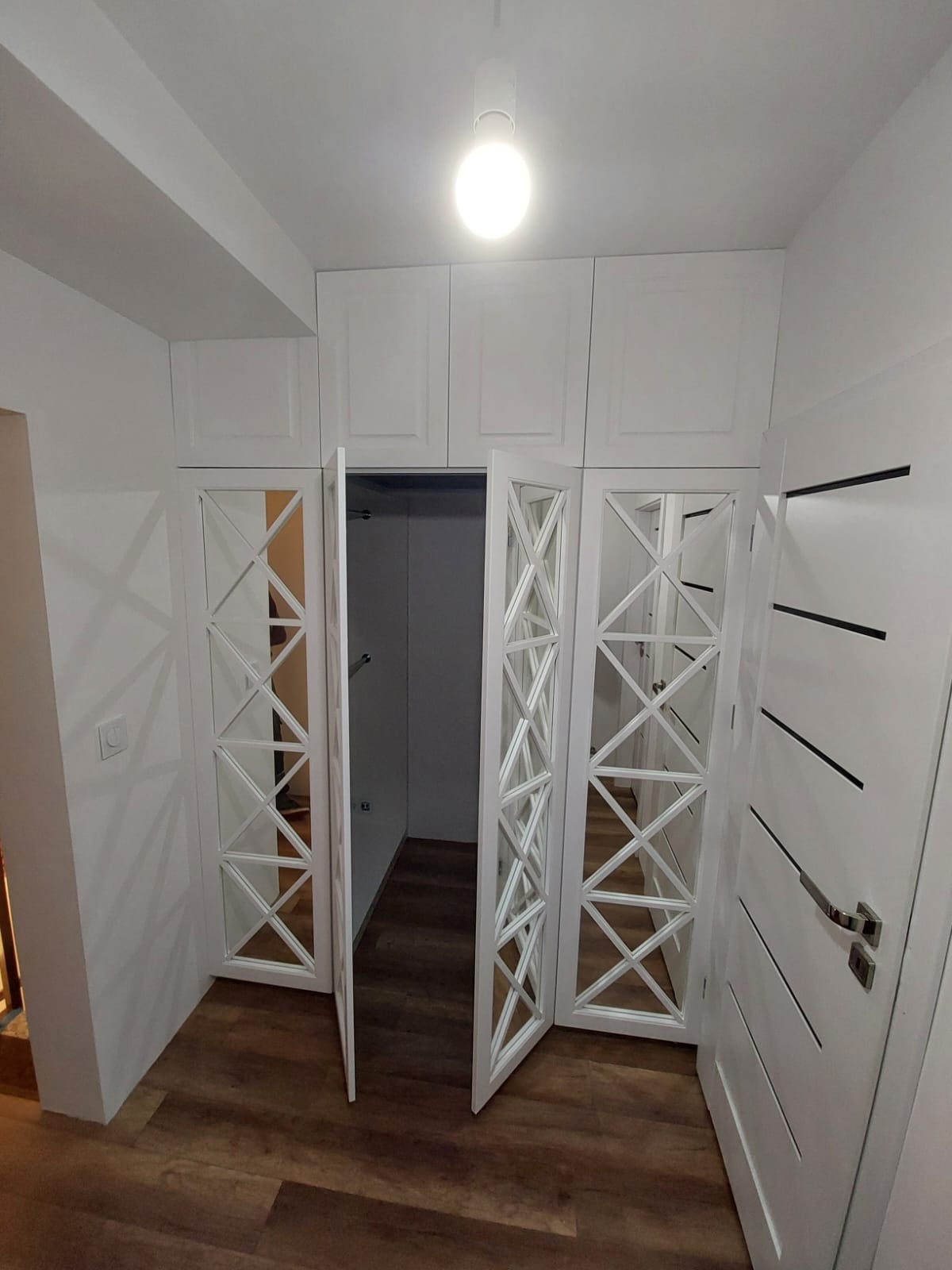 Biała szafa wnękowa z lustrzanymi drzwiami z geometrycznym wzorem, oświetlona lampą sufitową, widok z perspektywy podłogi, drewniana podłoga.