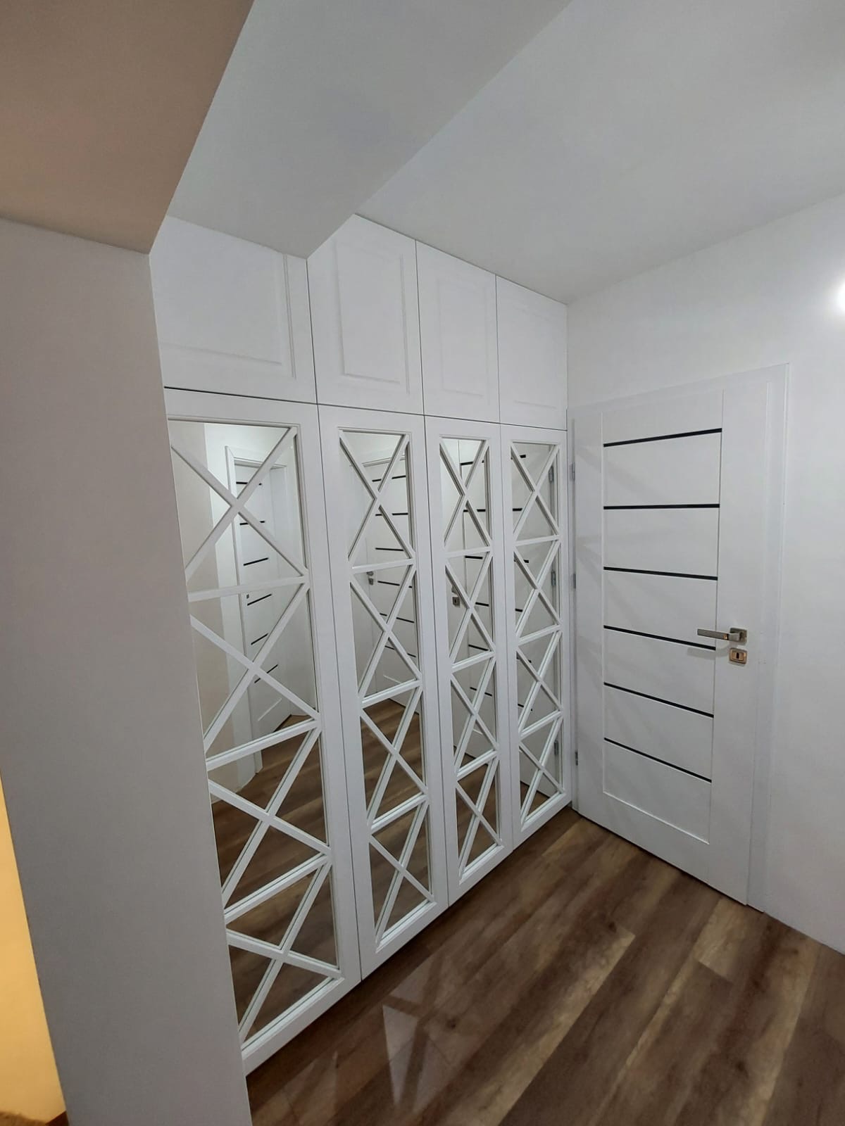 Biała szafa wnękowa z lustrami w geometryczne wzory, dopasowana do białych drzwi i drewnianej podłogi, tworząca spójną aranżację wnętrza.
