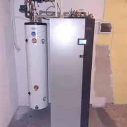 Zestaw grzewczy: pompa ciepła z zasobnikiem ciepłej wody użytkowej, widoczne rury instalacyjne i elektroniczny panel sterowania.