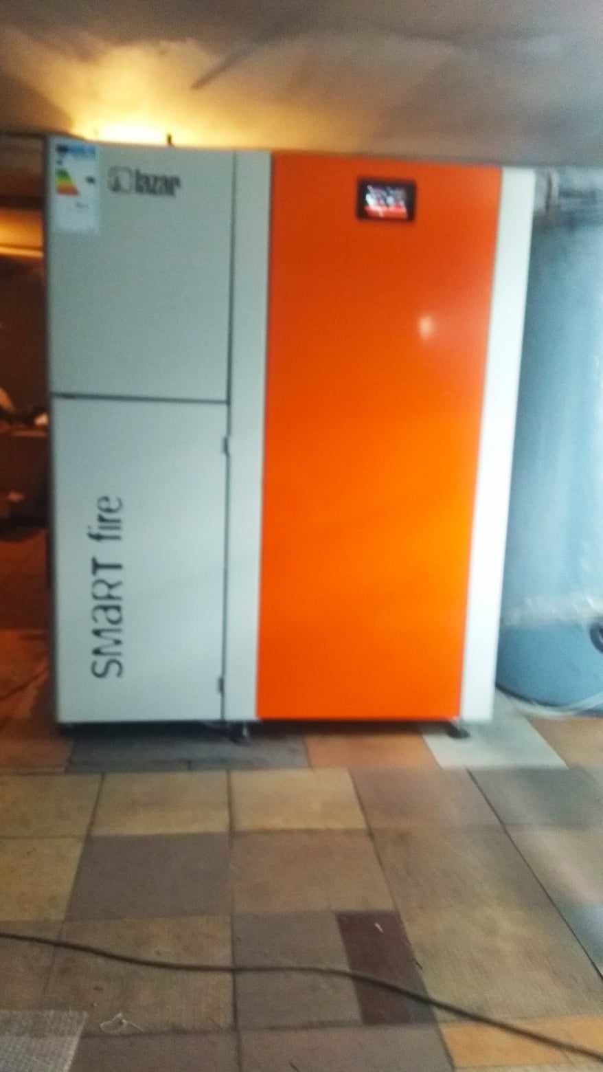 Nowoczesny piec CO Smart Fire Lazar z pomarańczowym panelem, zainstalowany w kotłowni z kafelkową podłogą.