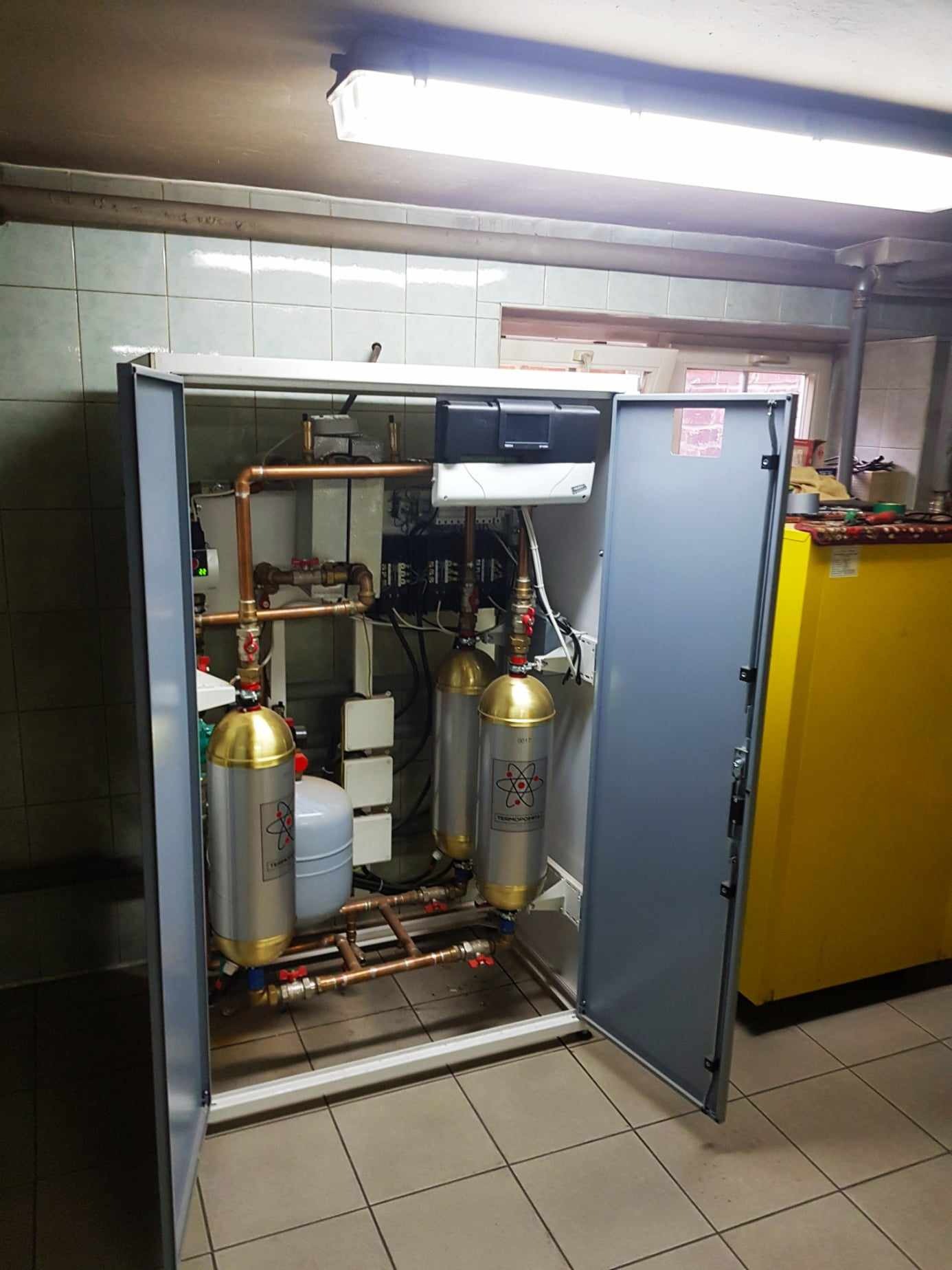 Otwarta szafka instalacyjna z zaworami, rurami miedzianymi i dwoma srebrnymi zbiornikami z złotymi nakładkami i logotypem atomu na tle ceramicznej ściany i podłogi.