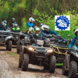 Grupa os&oacute;b w kaskach i strojach ochronnych na quadach, jadąca po błotnistej drodze w lesie, z logo 'Quady Pro' w prawym g&oacute;rnym rogu.