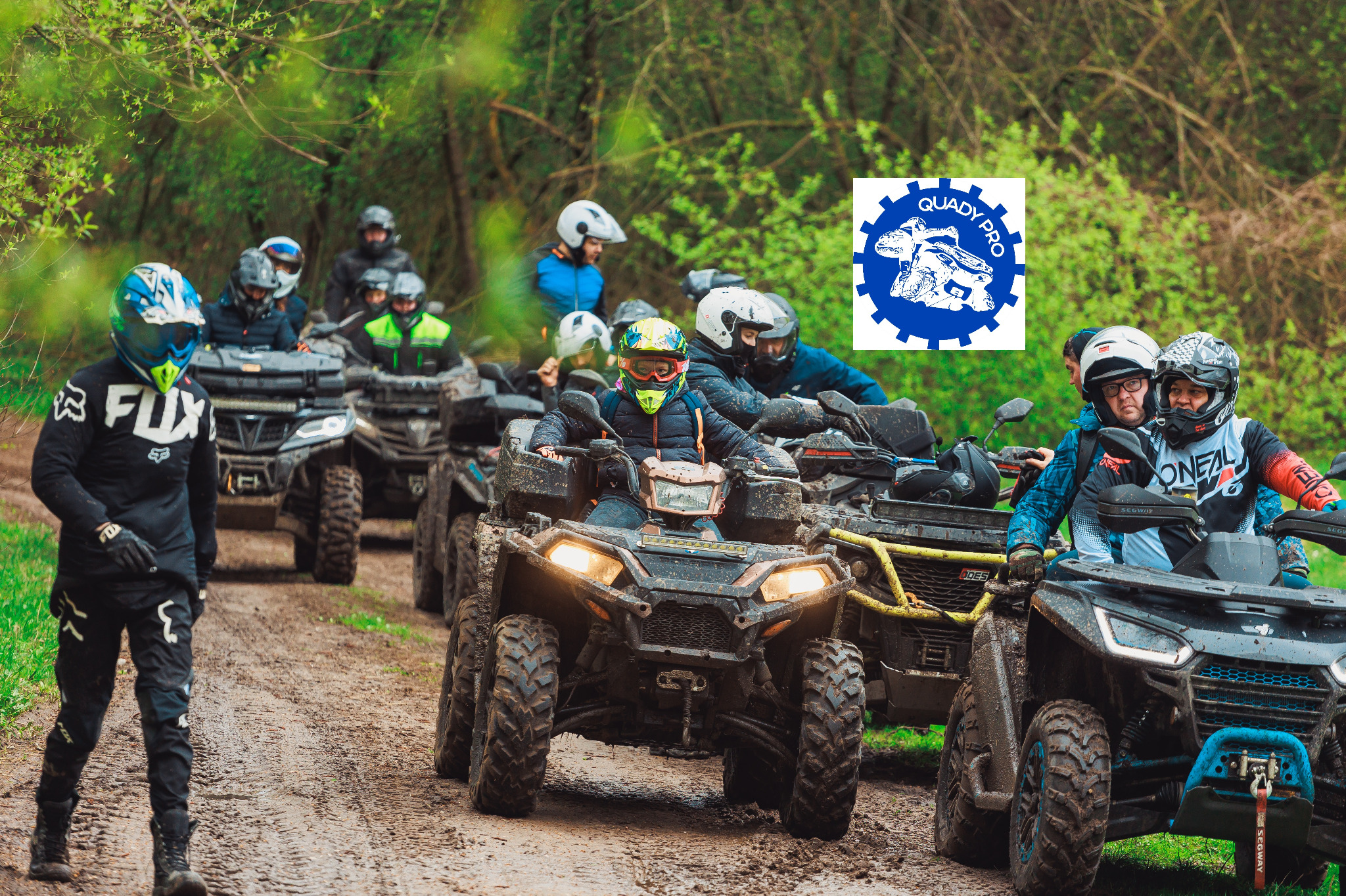 Grupa osób w kaskach i strojach ochronnych na quadach, jadąca po błotnistej drodze w lesie, z logo 'Quady Pro' w prawym górnym rogu.
