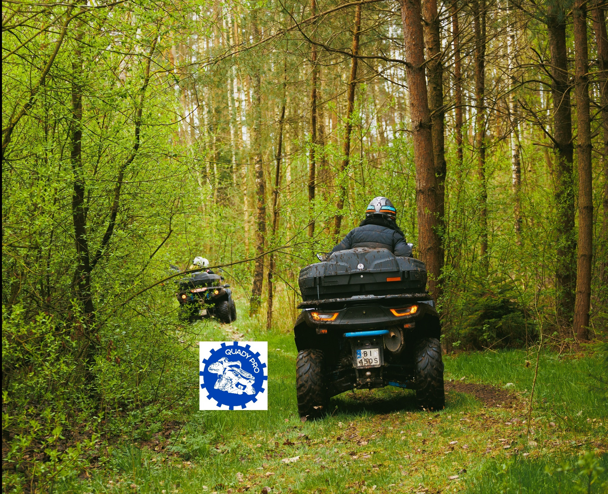 Dwa quady jadą leśną ścieżką, widok z tyłu na pierwszy pojazd z błotem na oponach i bagażnikiem, w tle drzewa i logo firmy Quady Pro w lewym dolnym rogu.