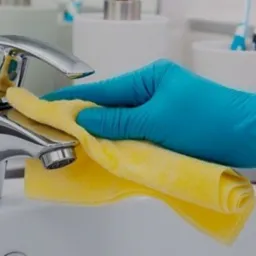 Błyszcząca chromowana bateria umywalkowa czyszczona żółtą ściereczką przez osobę w niebieskiej rękawicy ochronnej.