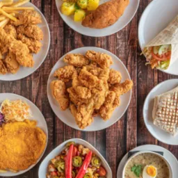 Widok z góry na stół z różnorodnym jedzeniem: panierowany kotlet z frytkami i surówkami, wrap z warzywami, zupa żurek z jajkiem, smażone kurczaki na talerzach, sałatka z czerwoną papryką...