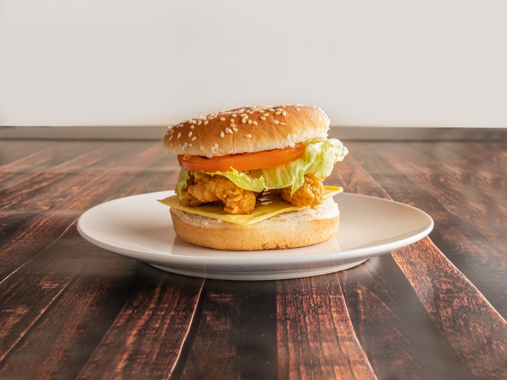 Burger z panierowanym kurczakiem, serem, pomidorem i sałatą na białym talerzu, ustawiony na drewnianym blacie.