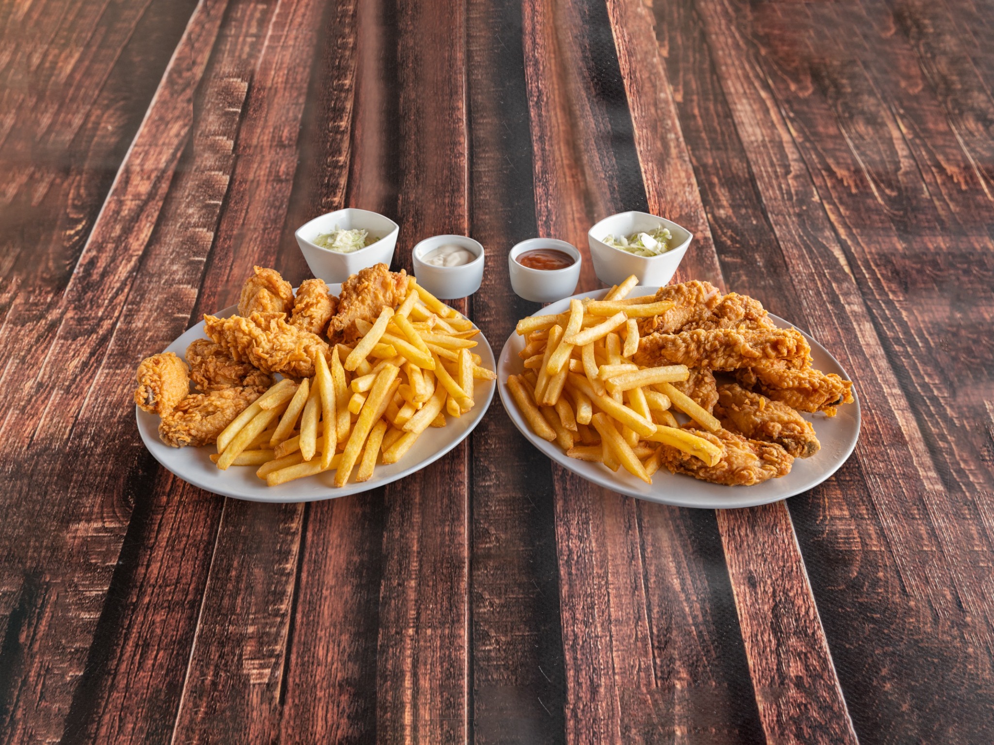 Dwa talerze z frytkami i kurczakiem w panierce, uzupełnione o sosy i surówkę colesław, zaprezentowane na drewnianym stole.