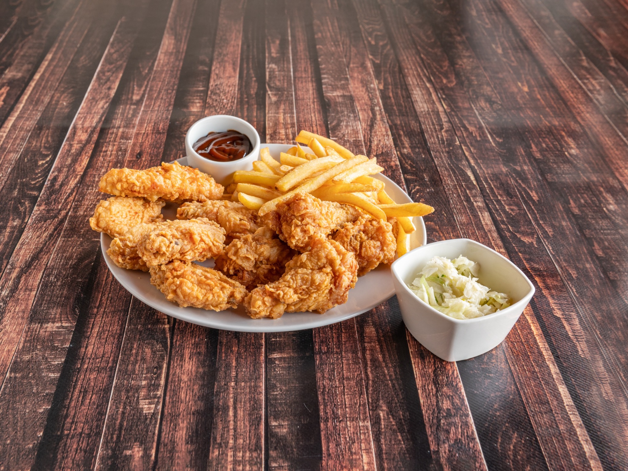 Porcja panierowanych skrzydełek kurczaka z frytkami, sosem barbecue i surówką colesław, podana na białym talerzu na ciemnym, drewnianym stole.