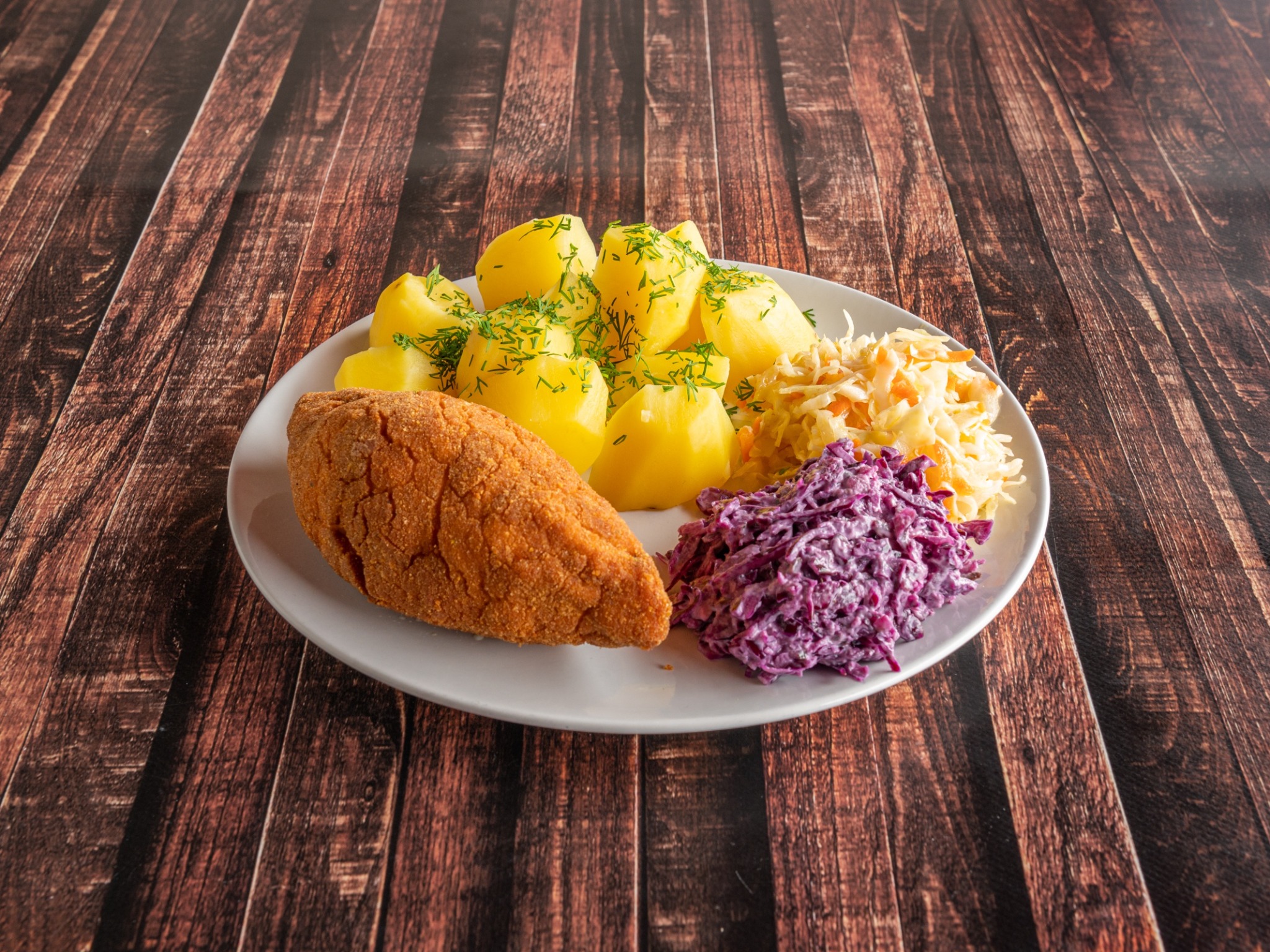 Kotlet schabowy w panierce, gotowane ziemniaki posypane koperkiem oraz dwie surówki na białym talerzu, na tle drewnianego blatu.