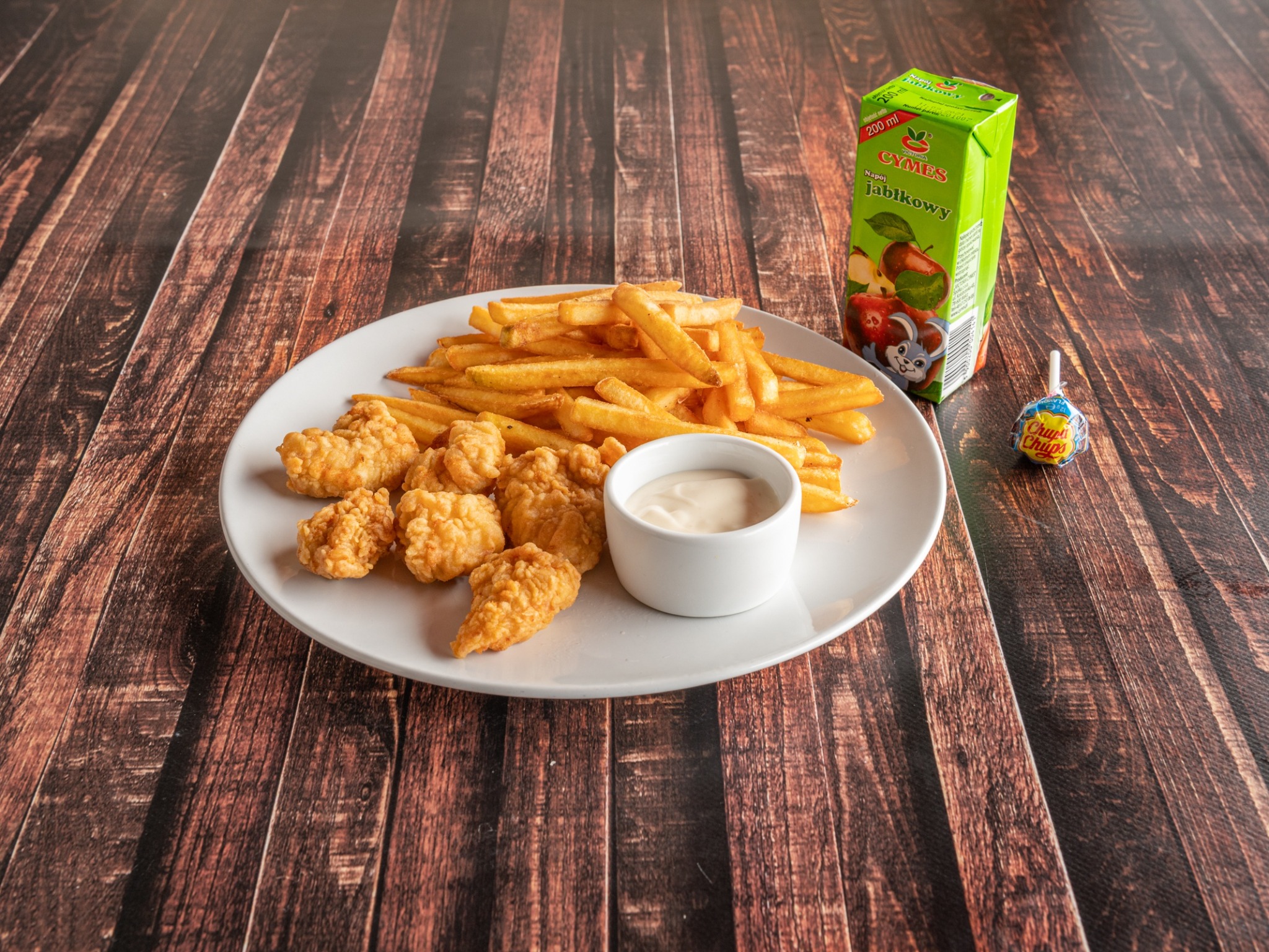 Frytki, nuggets z kurczaka i dip w białej miseczce na białym talerzu, obok kartonik soku jabłkowego i lizak Chupa Chups, na drewnianym stole.