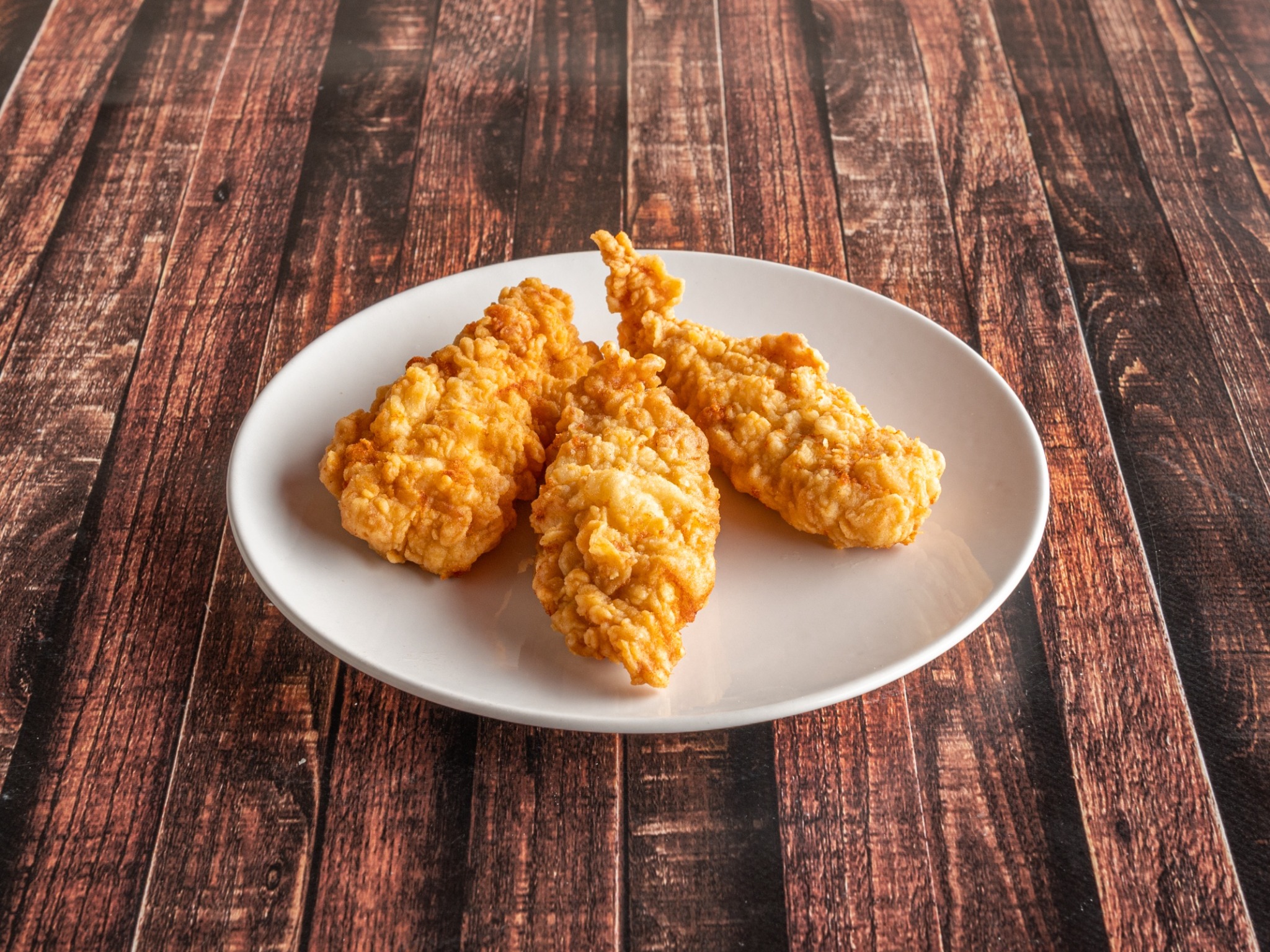 Trzy chrupiące, złociste filety z kurczaka w panierce na białym talerzu, ułożone na drewnianym stole z widoczną fakturą słojów.