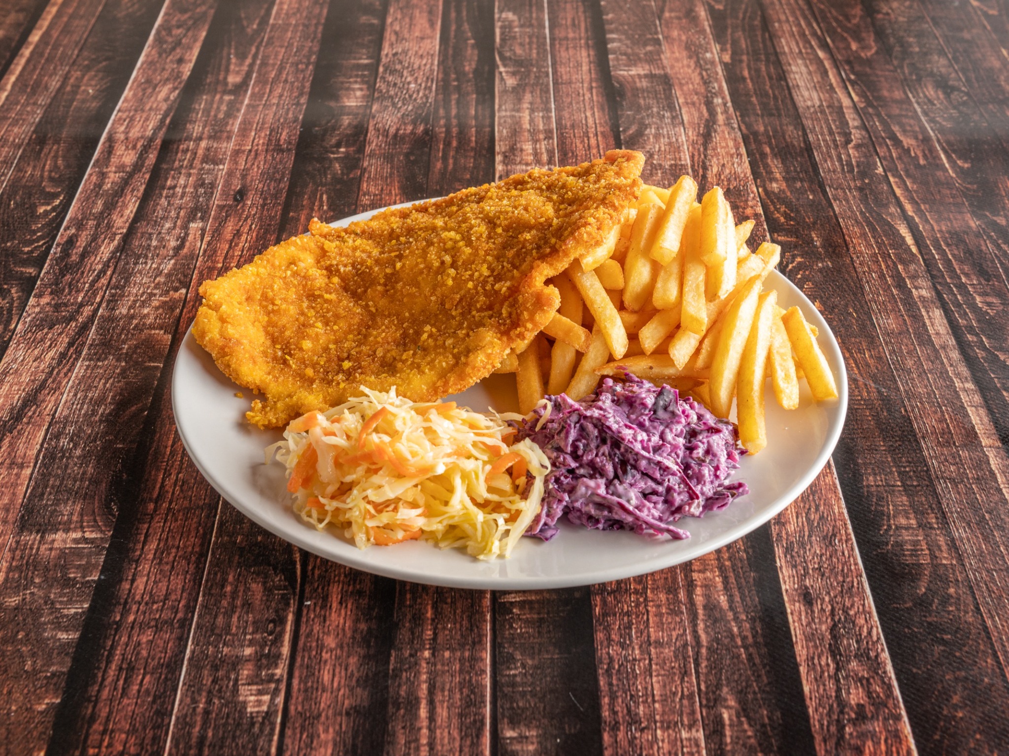 Zestaw obiadowy: panierowana ryba, frytki i dwie sałatki colesław (biała i czerwona) na białym talerzu, ułożone na ciemnym drewnianym stole.