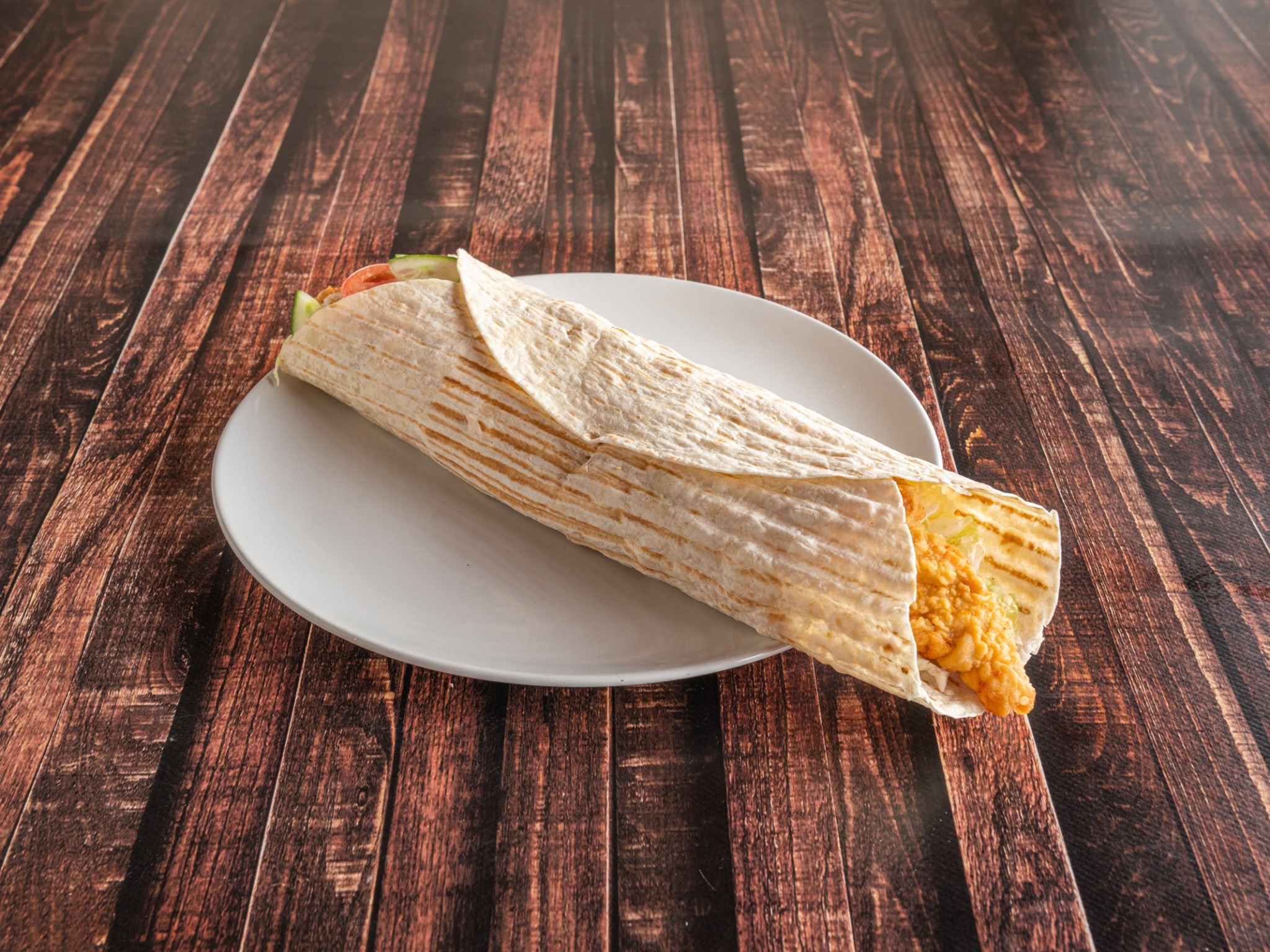 Tortilla wrap z panierowanym kurczakiem, sałatą, pomidorem i ogórkiem na białym talerzu, ułożona na ciemnym drewnianym stole.