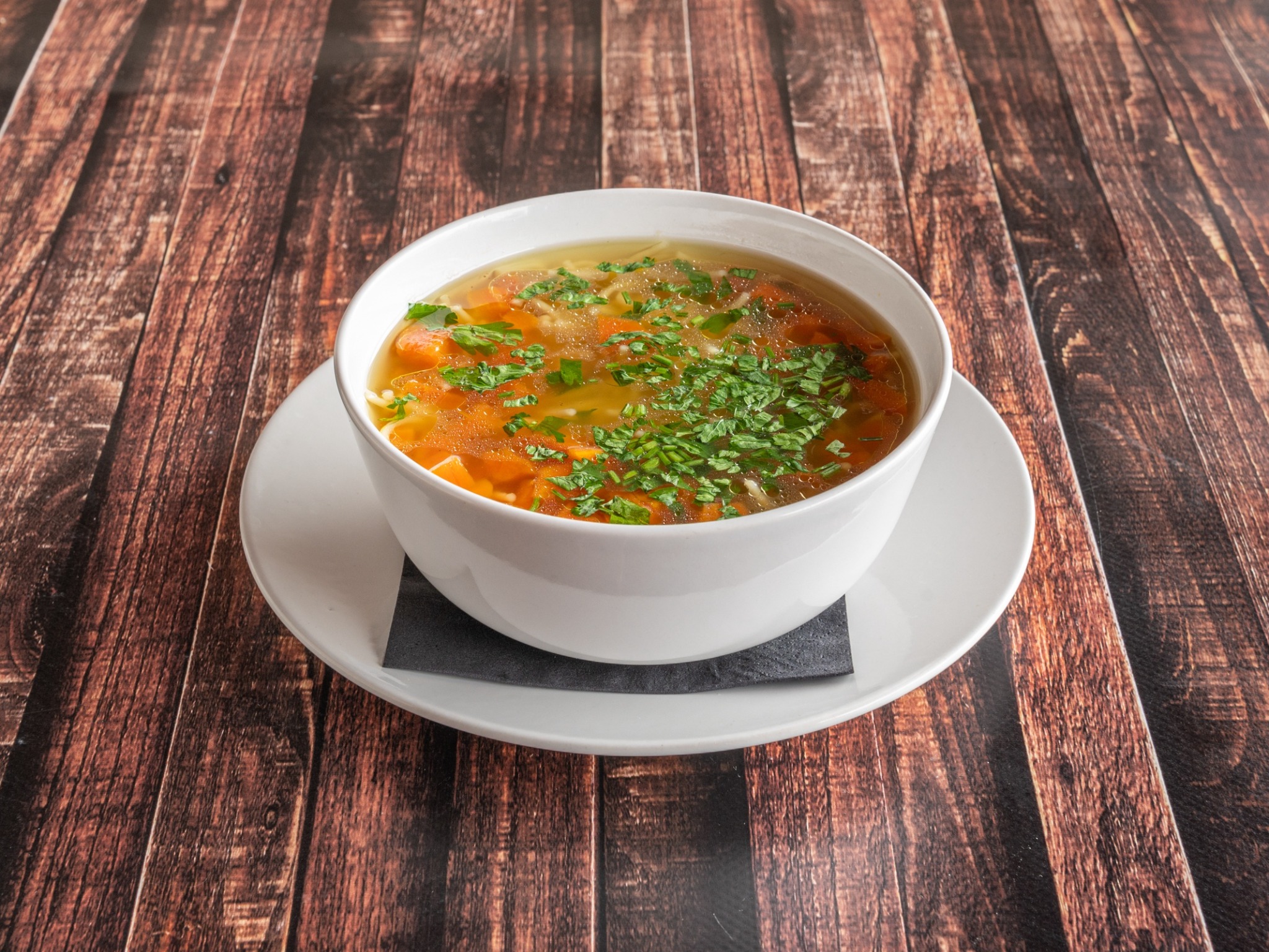 Apetyczny bulion z marchewką, makaronem i posiekaną natką pietruszki, podany w białej miseczce na drewnianym stole.
