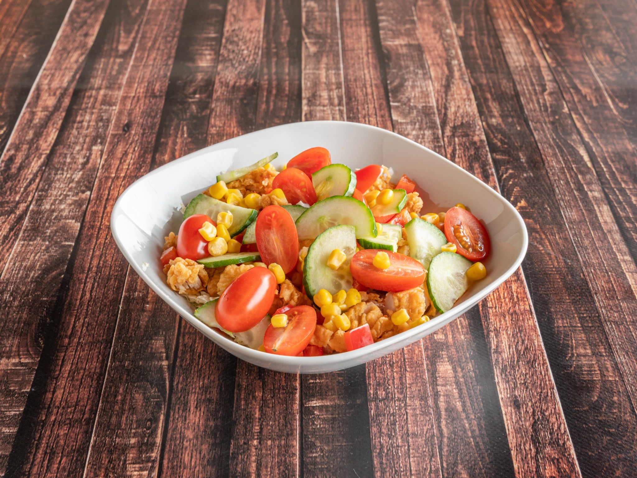 Sałatka z kurczakiem, pomidorkami cherry, ogórkiem i kukurydzą w białej misce na drewnianym stole.