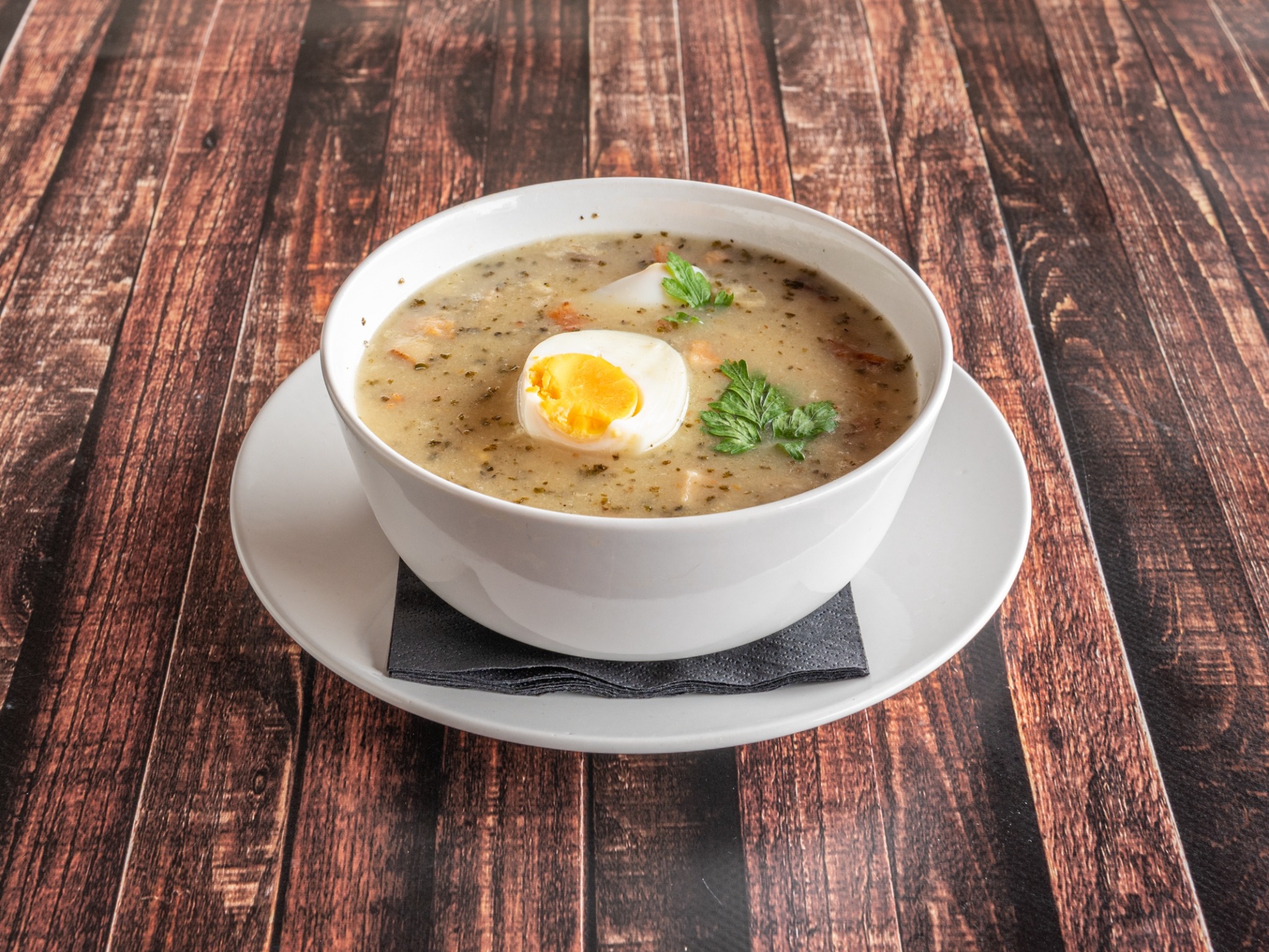 Żurek w białej misce z połówką jajka i natką pietruszki, podany na białym talerzu z szarą serwetką na drewnianym stole.