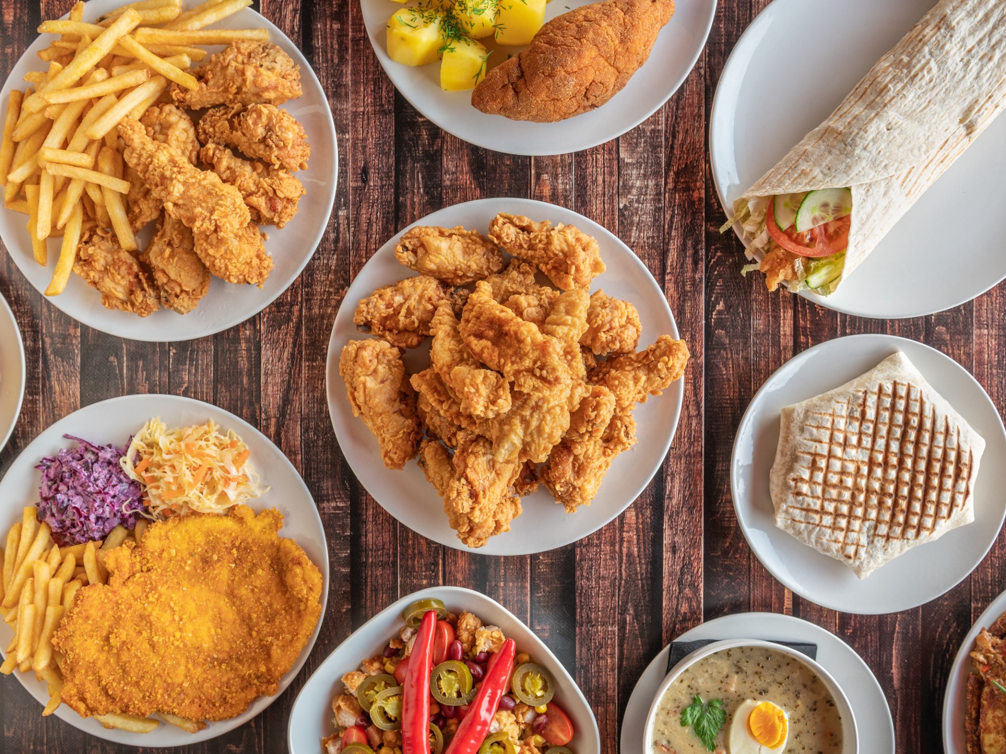 Widok z góry na stół z różnorodnym jedzeniem: panierowany kotlet z frytkami i surówkami, wrap z warzywami, zupa żurek z jajkiem, smażone kurczaki na talerzach, sałatka z czerwoną papryką...