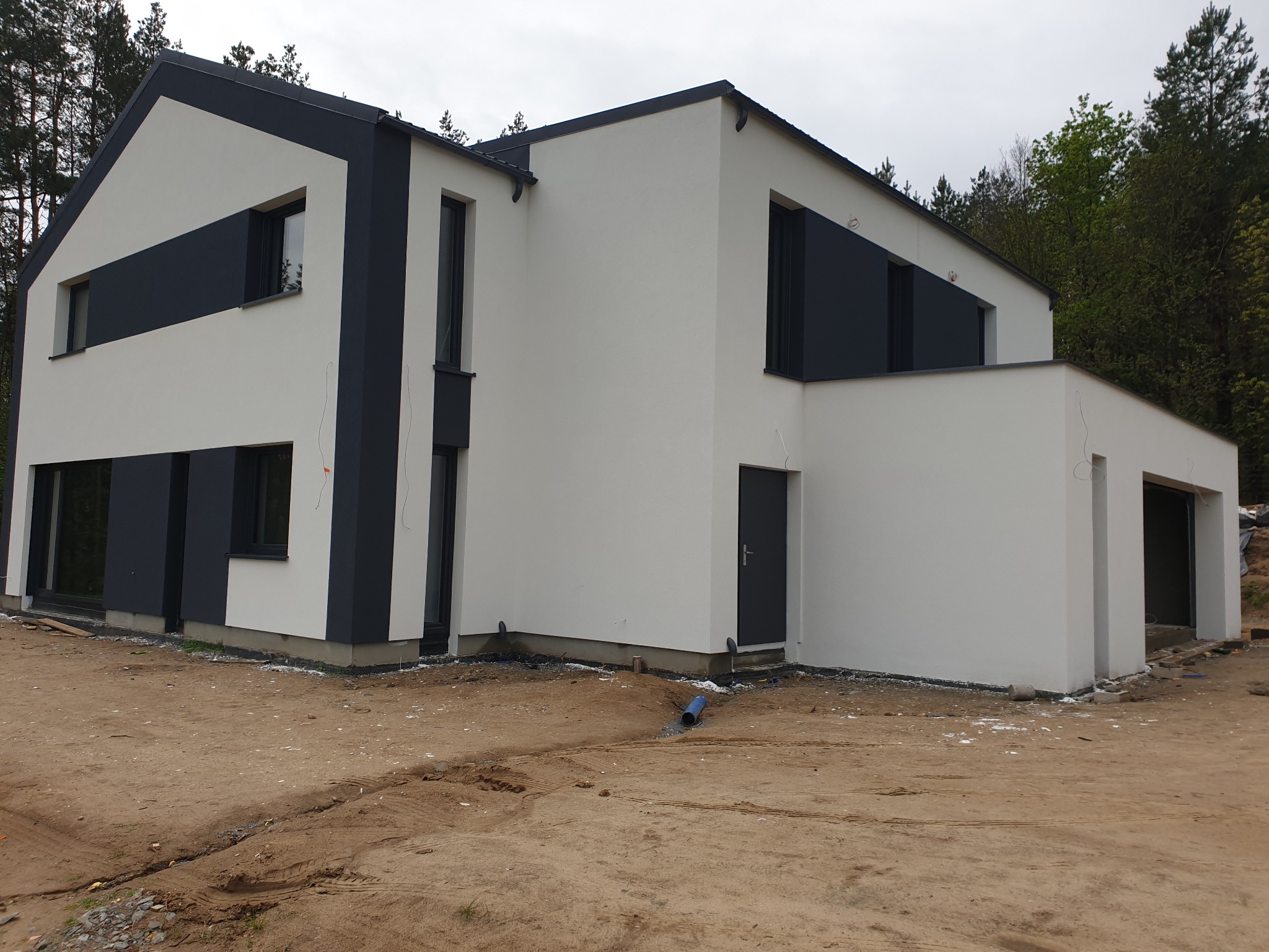 Nowoczesny dom jednorodzinny z białą elewacją i grafitowymi akcentami, widoczny z poziomu gruntu, teren wokół w trakcie zagospodarowywania, na pierwszym planie rura odprowadzająca wodę.