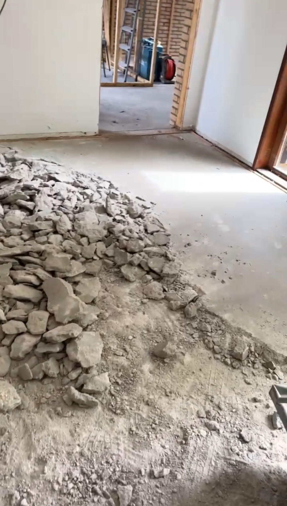 Sterta gruzu po rozbiórce podłogi wewnątrz pomieszczenia; widoczne fragmenty betonu i zaprawy, w tle odsłonięta konstrukcja ściany z cegły i drewnianych belek.